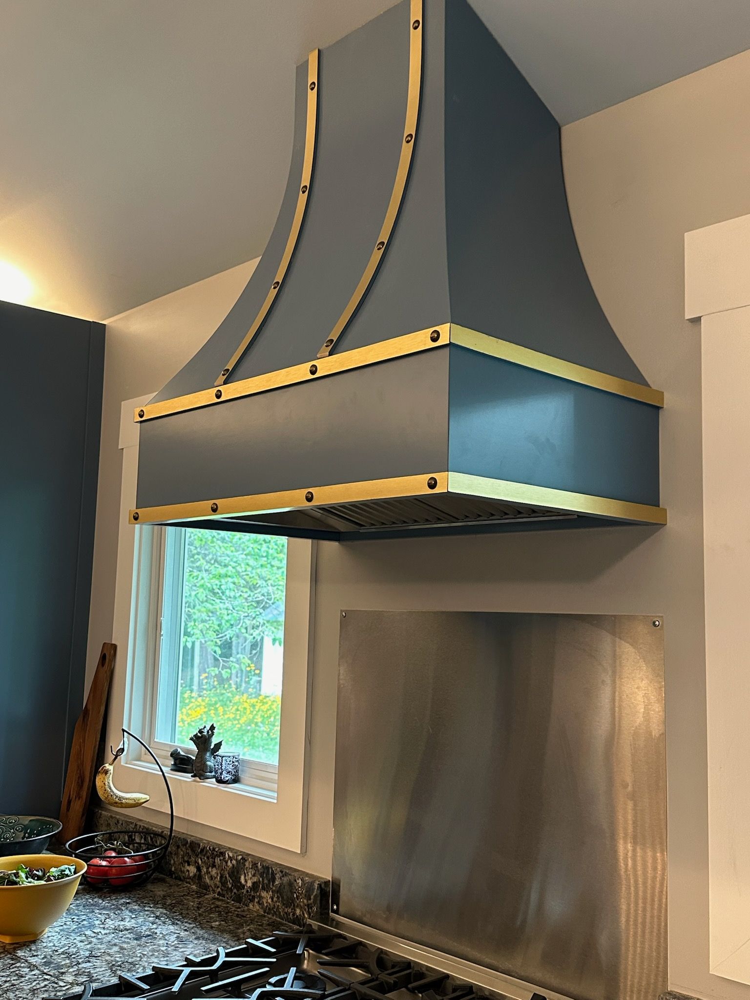 Kitchen range hood with blue-gray finish and gold trim above a stove area, beside a window and backsplash