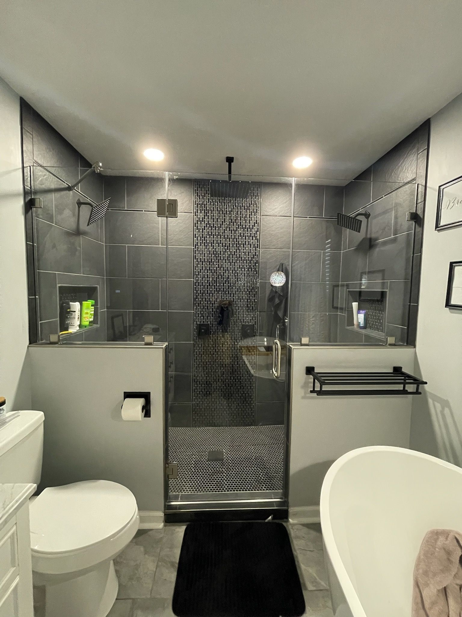 Modern black-tiled bathroom with toilet, shower stall, sink, and black rug