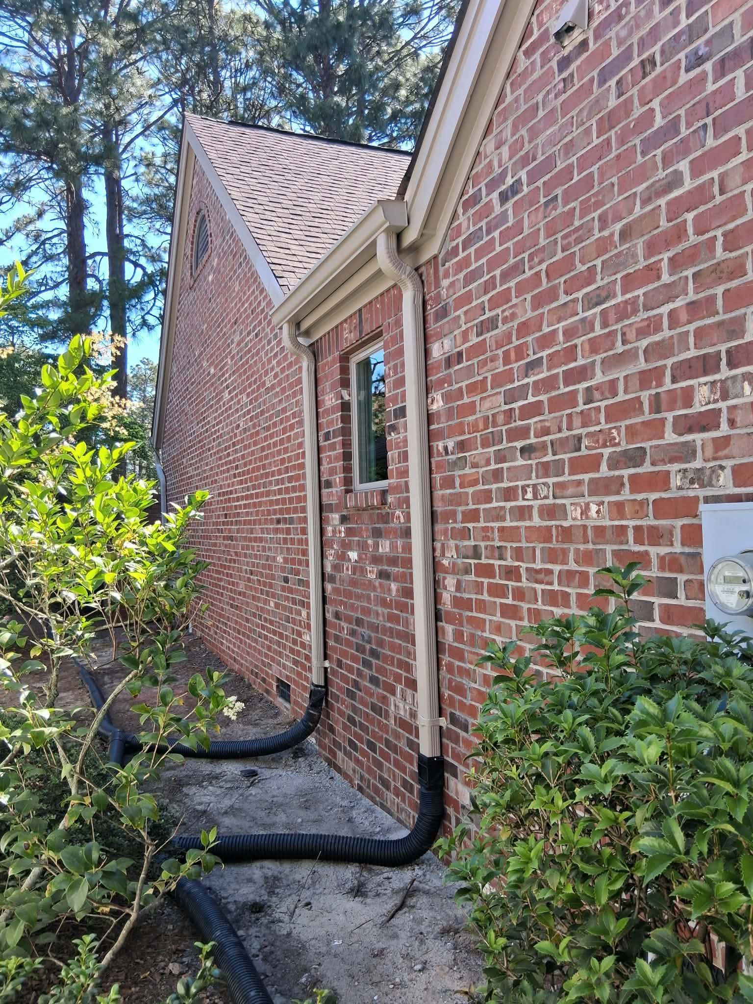 Brick house side with narrow walkway, black downspouts, and shrubs in a wooded yard.