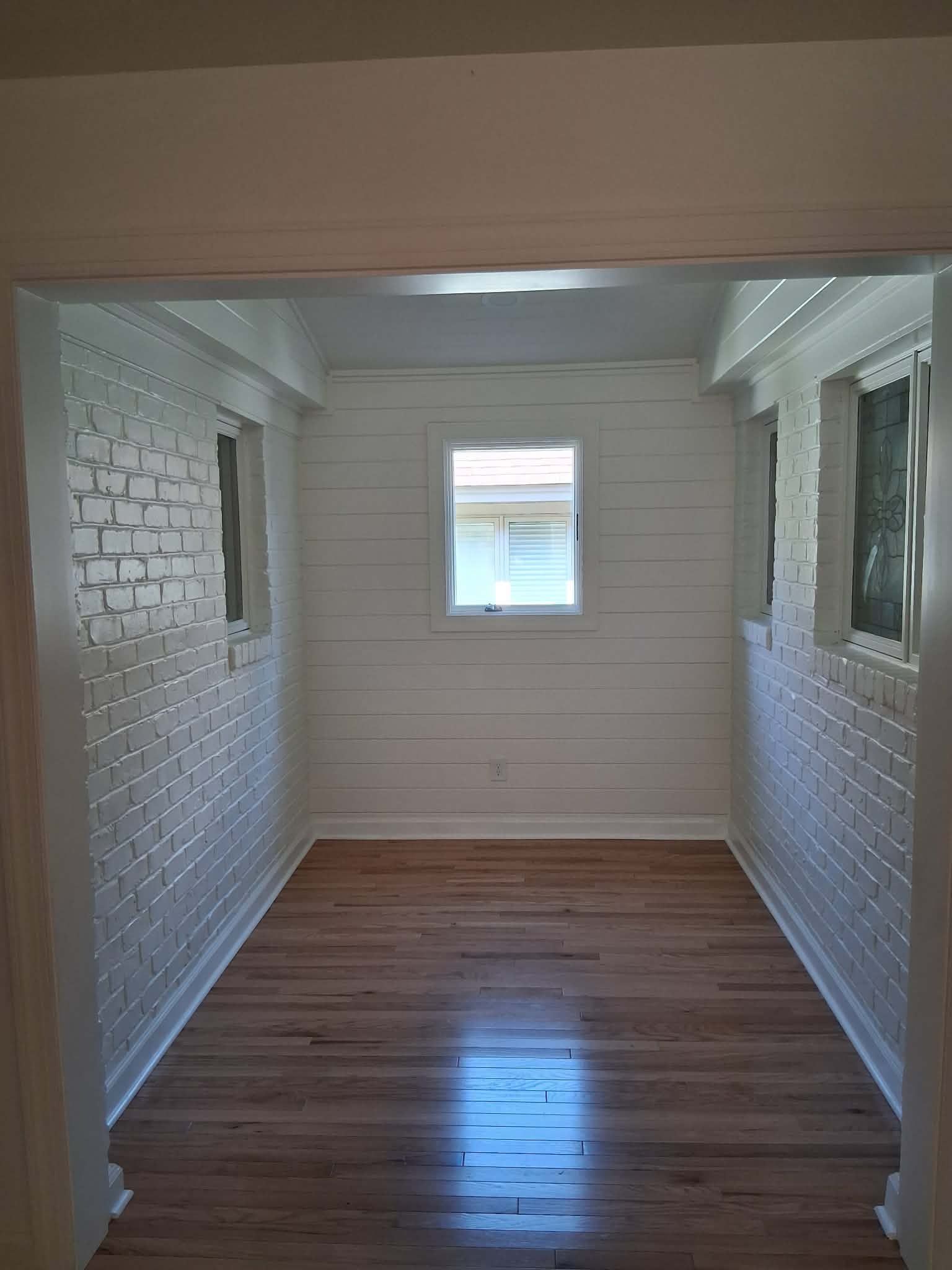 Narrow empty room with white brick walls, wood floor, and two windows at the far end.