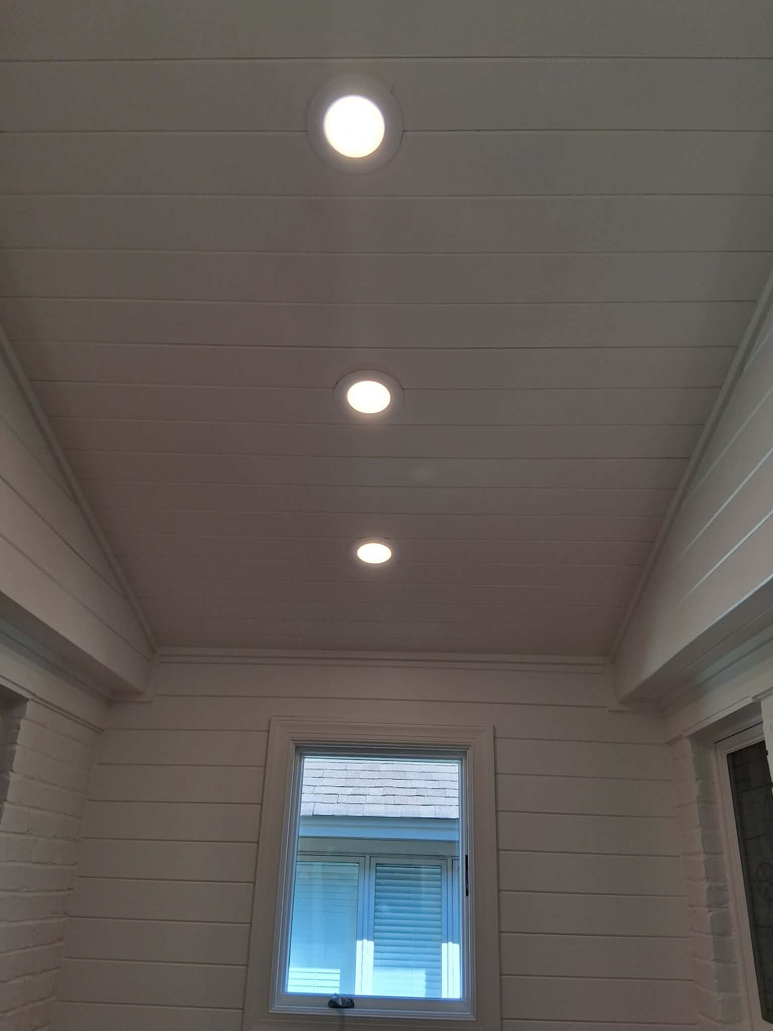 Covered porch ceiling with three lit recessed lights and a window at the end.