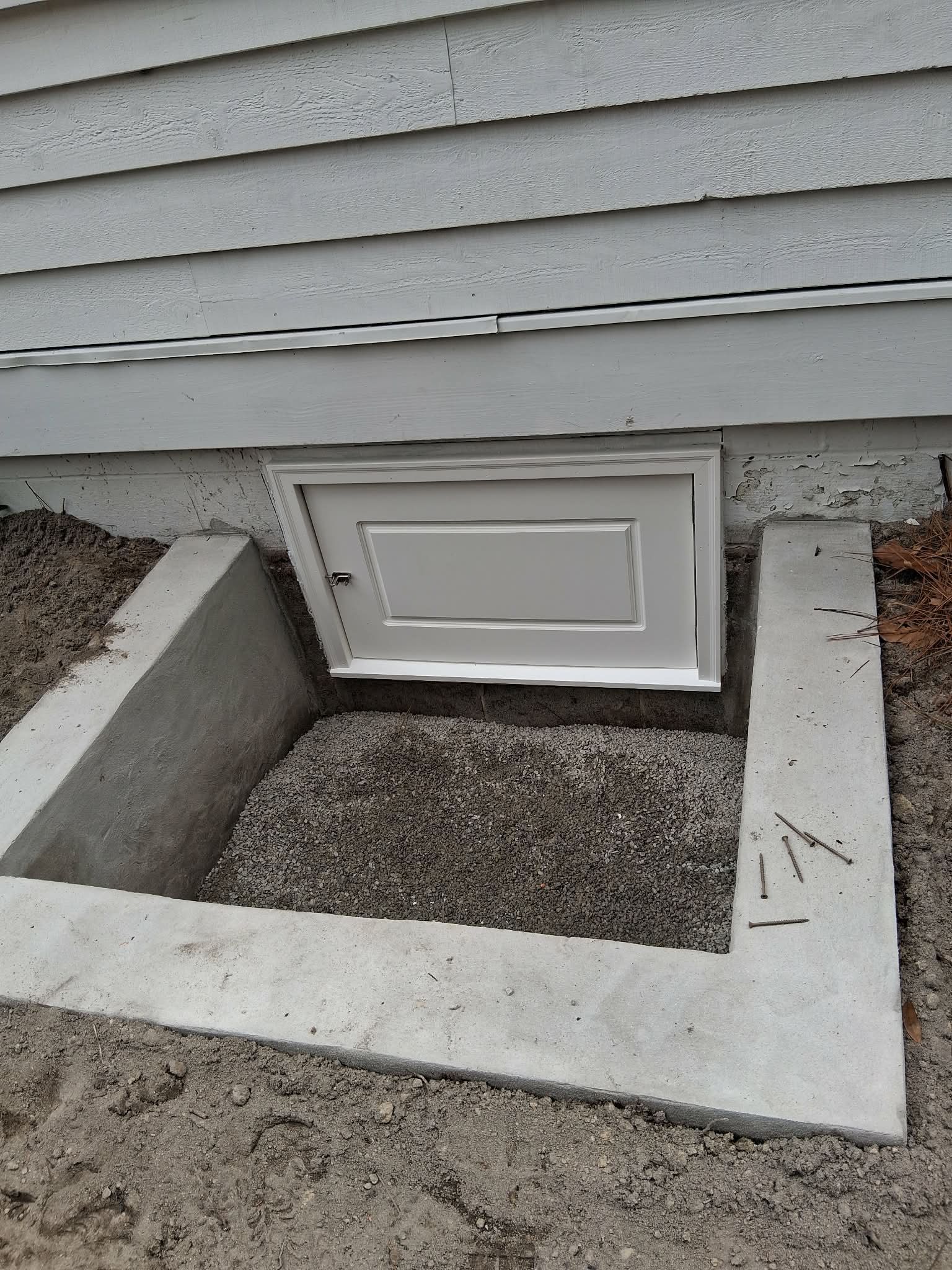Small basement window well with a white access door under house siding