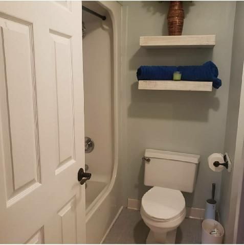 Small bathroom with toilet, tub, shelves, and towels; beige walls and white fixtures.