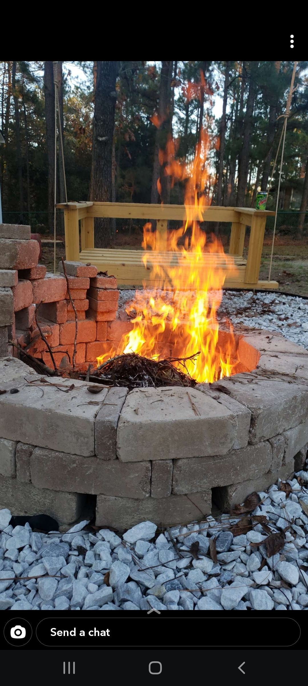 Fire burning in a circular brick fire pit outdoors, with trees and a wooden bench in the background.