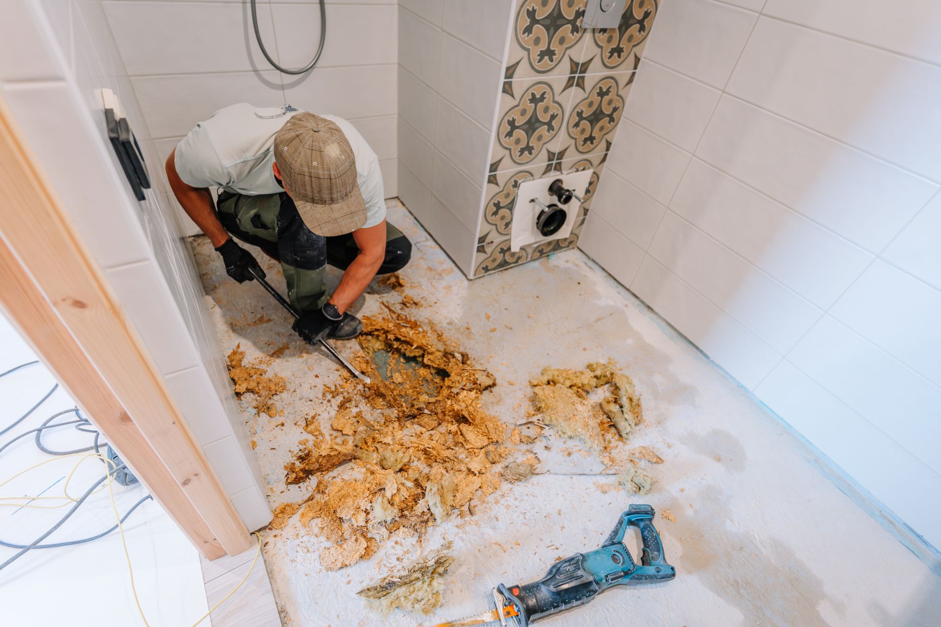 Worker cleaning a bathroom floor covered in debris and mud, with a drill on the ground