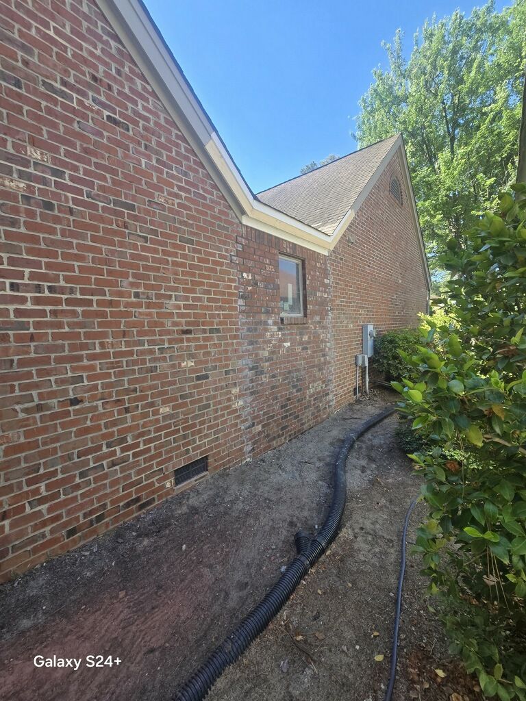 Brick house side yard with narrow gravel path, downspout, and bushes beside the exterior wall