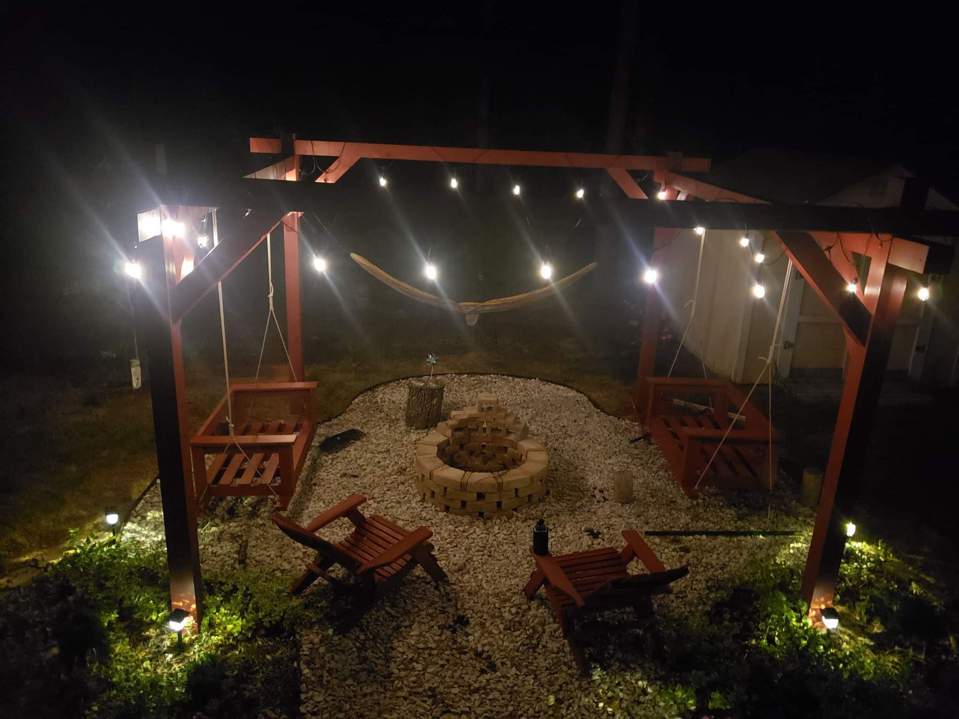 Nighttime outdoor fire pit area under string lights, with wooden seating and gravel ground.