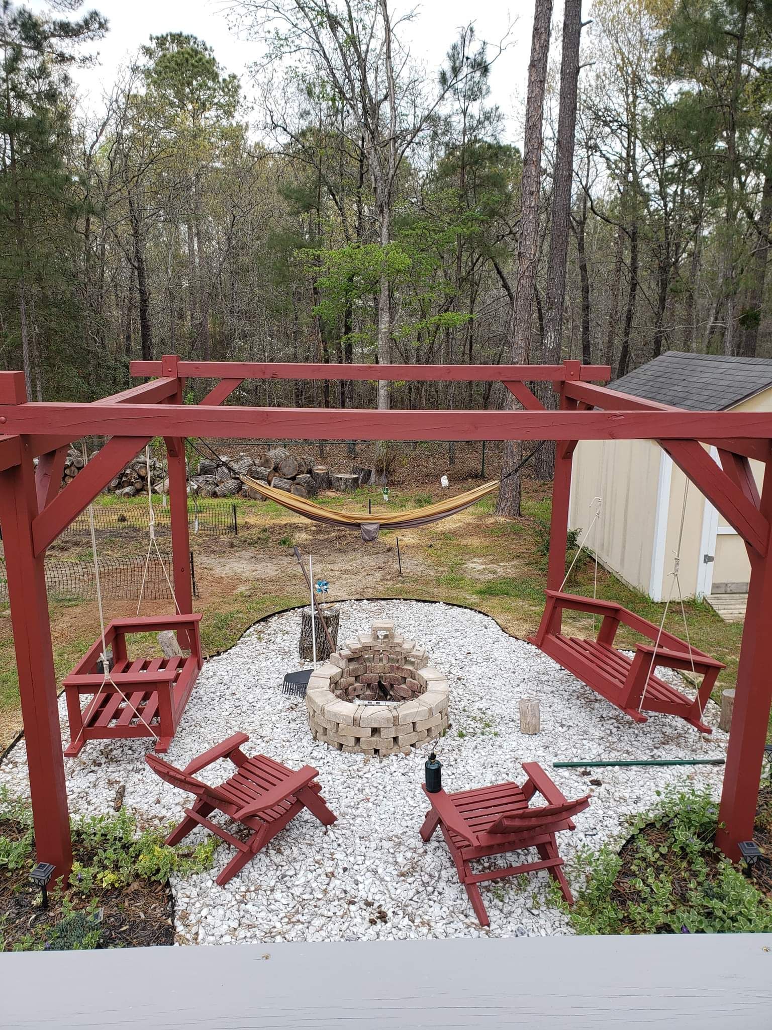 Backyard fire pit area with red pergola, white gravel, and Adirondack chairs in a wooded setting