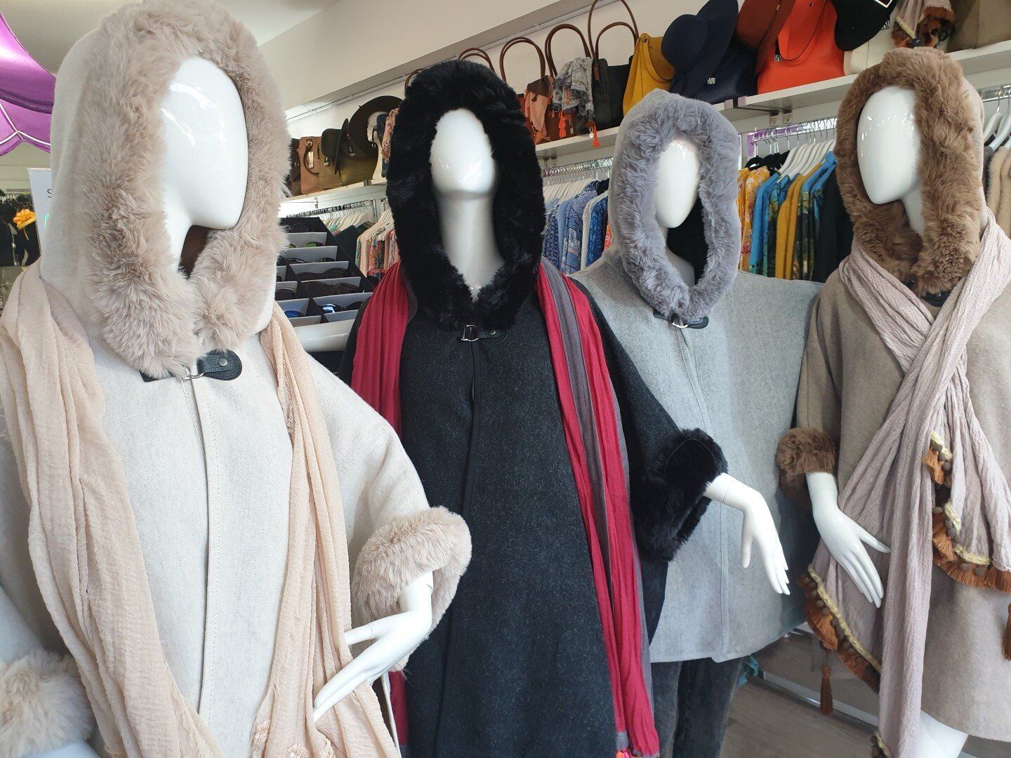 Four mannequins wearing hooded capes in a store. The capes are beige, black, grey and brown.