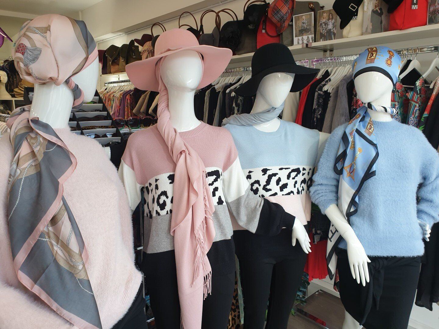 Four mannequins wearing fashionable sweaters, hats, scarves, and black pants in a clothing store.