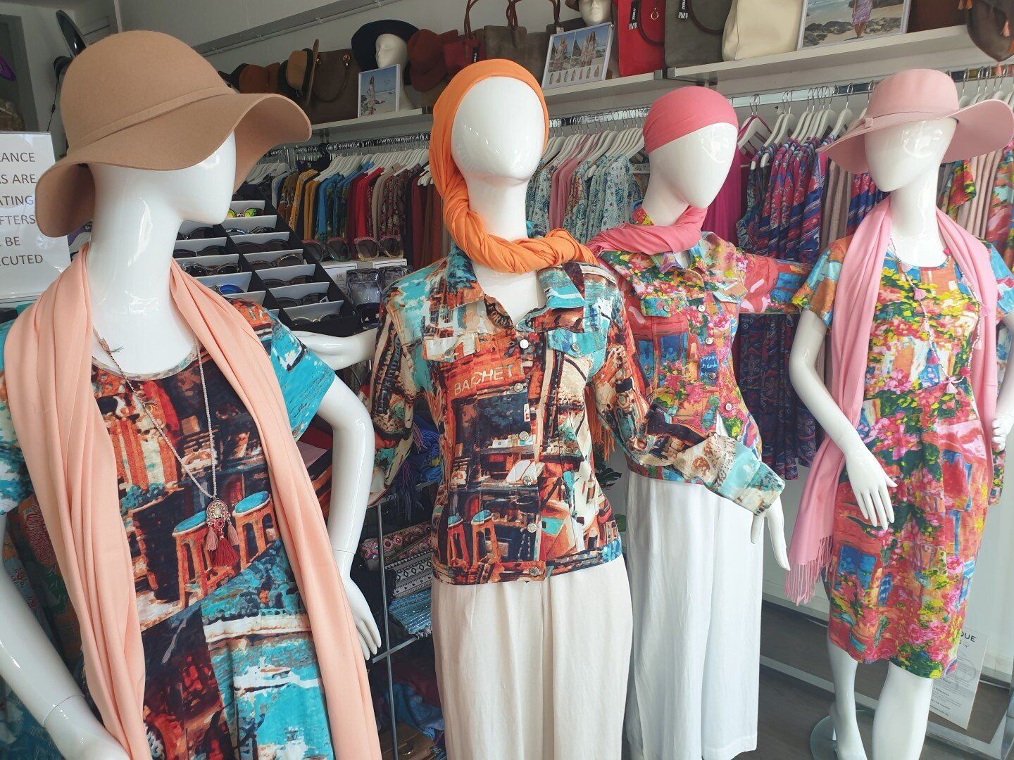 Four mannequins in a store window wearing colorful printed outfits, scarves, and hats.