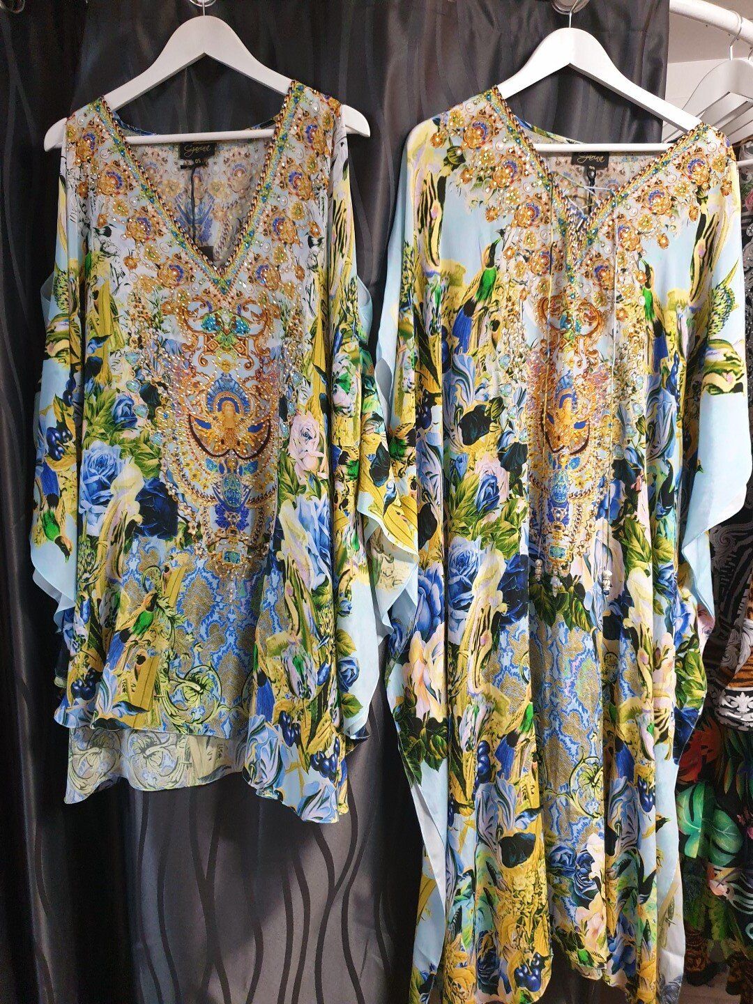 Two colorful, patterned kaftans on hangers against a dark background. One is cold-shoulder style.