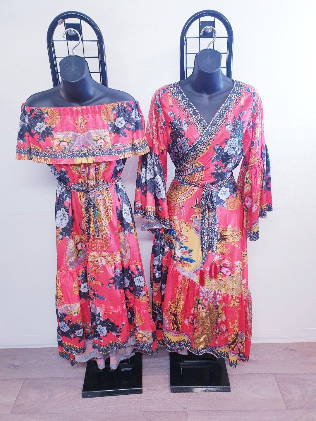 Two floral print dresses on black mannequins; one off-shoulder and one wrap style; both with belts; red background.