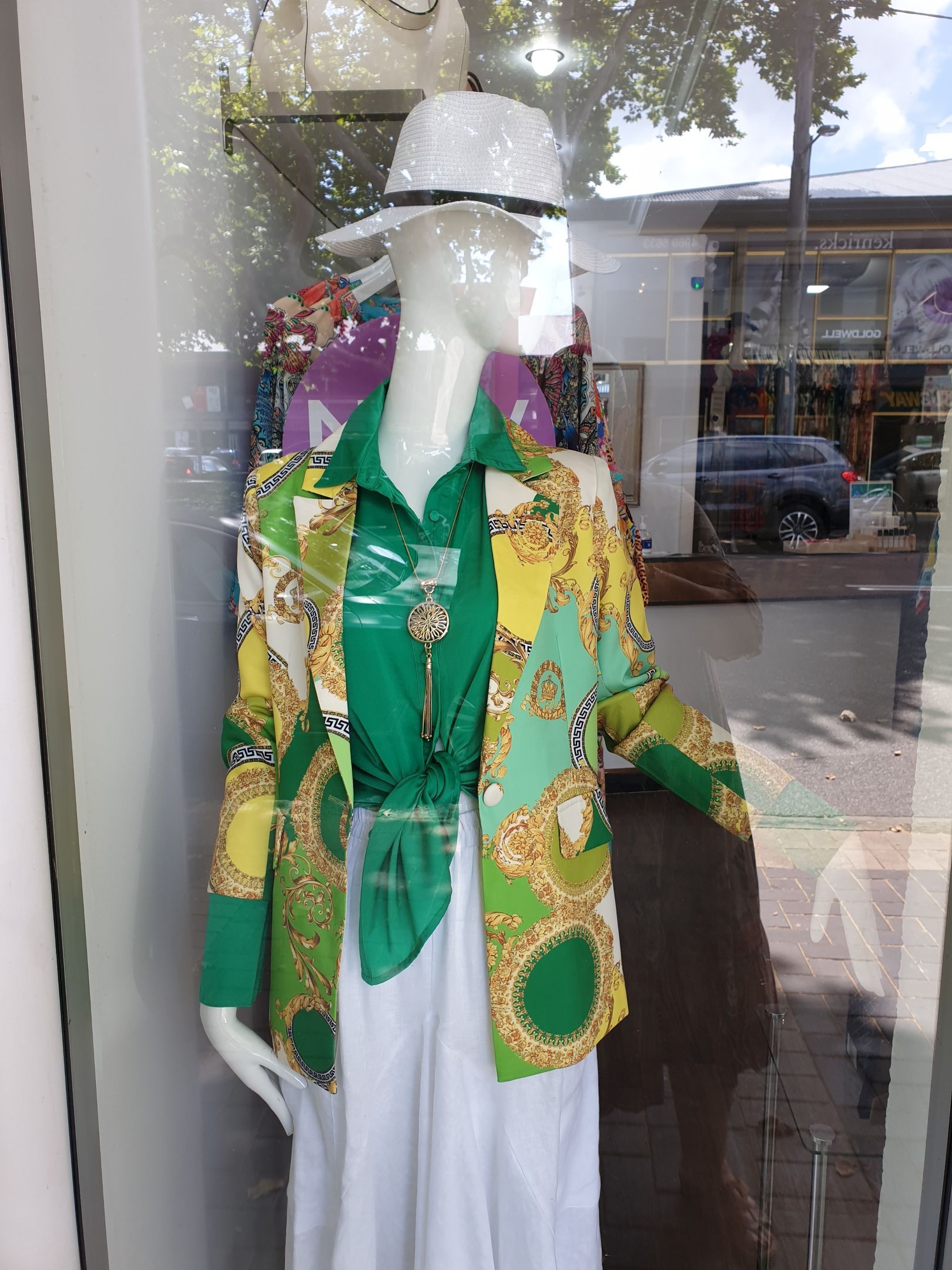 Mannequin in shop window wearing a green and yellow patterned blazer, green top, white pants, and a white hat.
