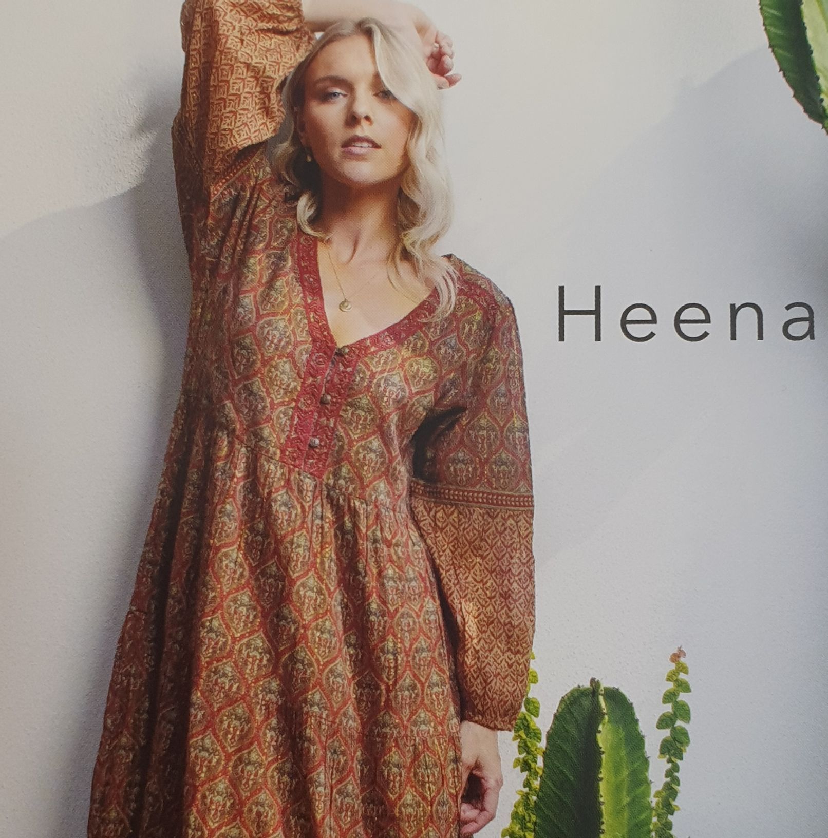 Woman wearing patterned brown dress, leaning against wall, arm raised.
