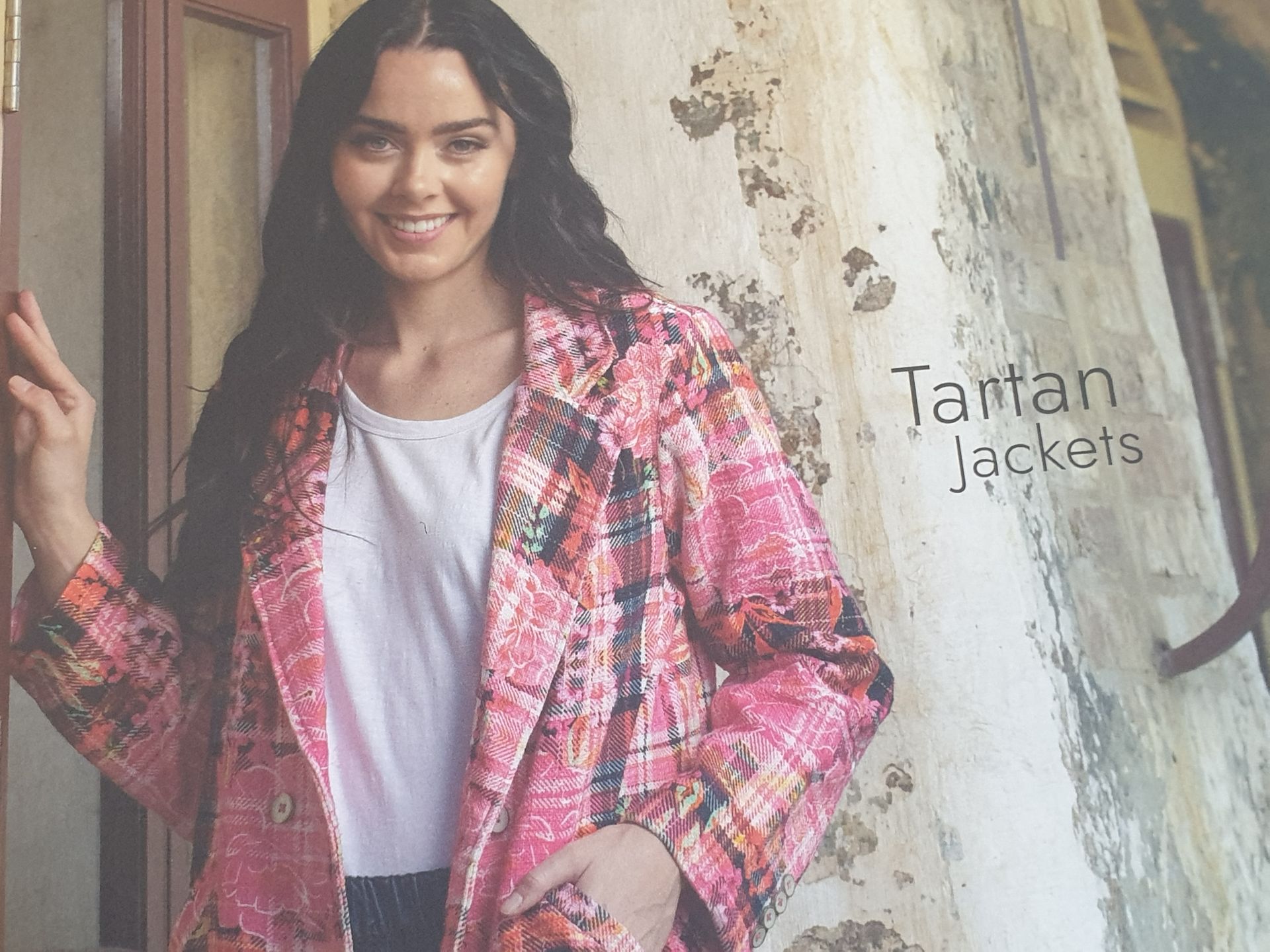 Woman in pink patterned jacket, smiling. Leaning on a stone wall, outdoor setting.