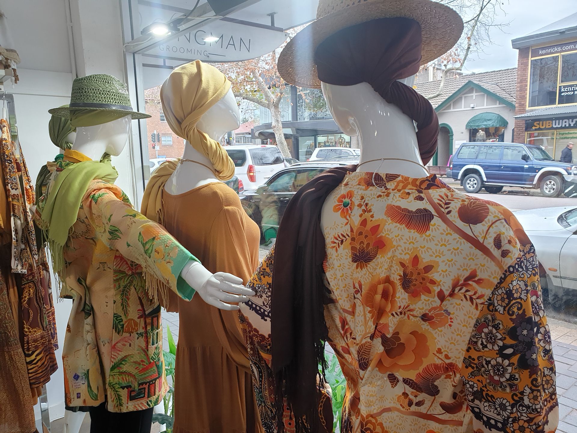 Three mannequins dressed in patterned clothing inside a shop window, with a street scene visible in the background.