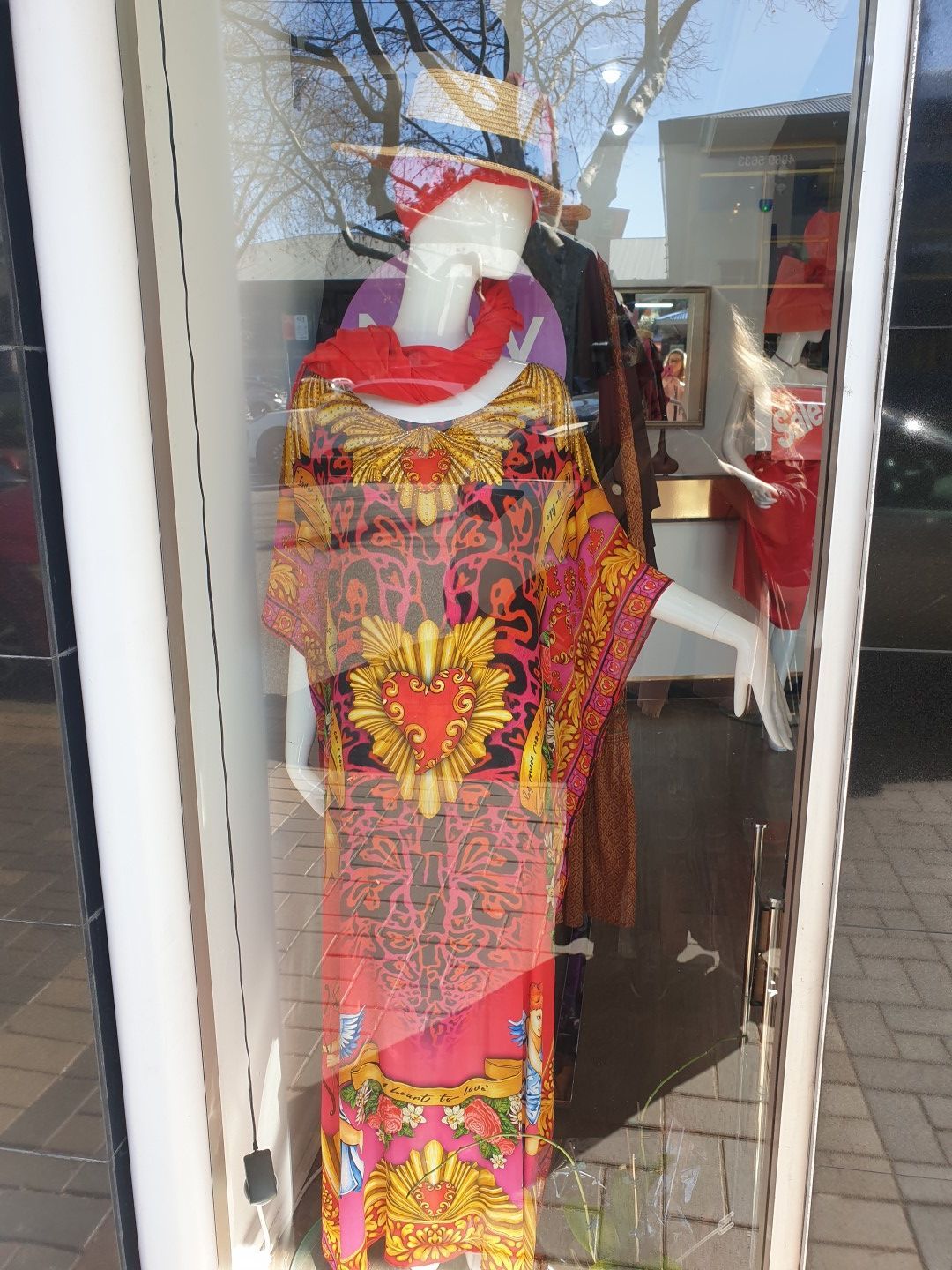 Mannequin in a shop window wearing a vibrant pink and gold patterned dress, red scarf, and hat.