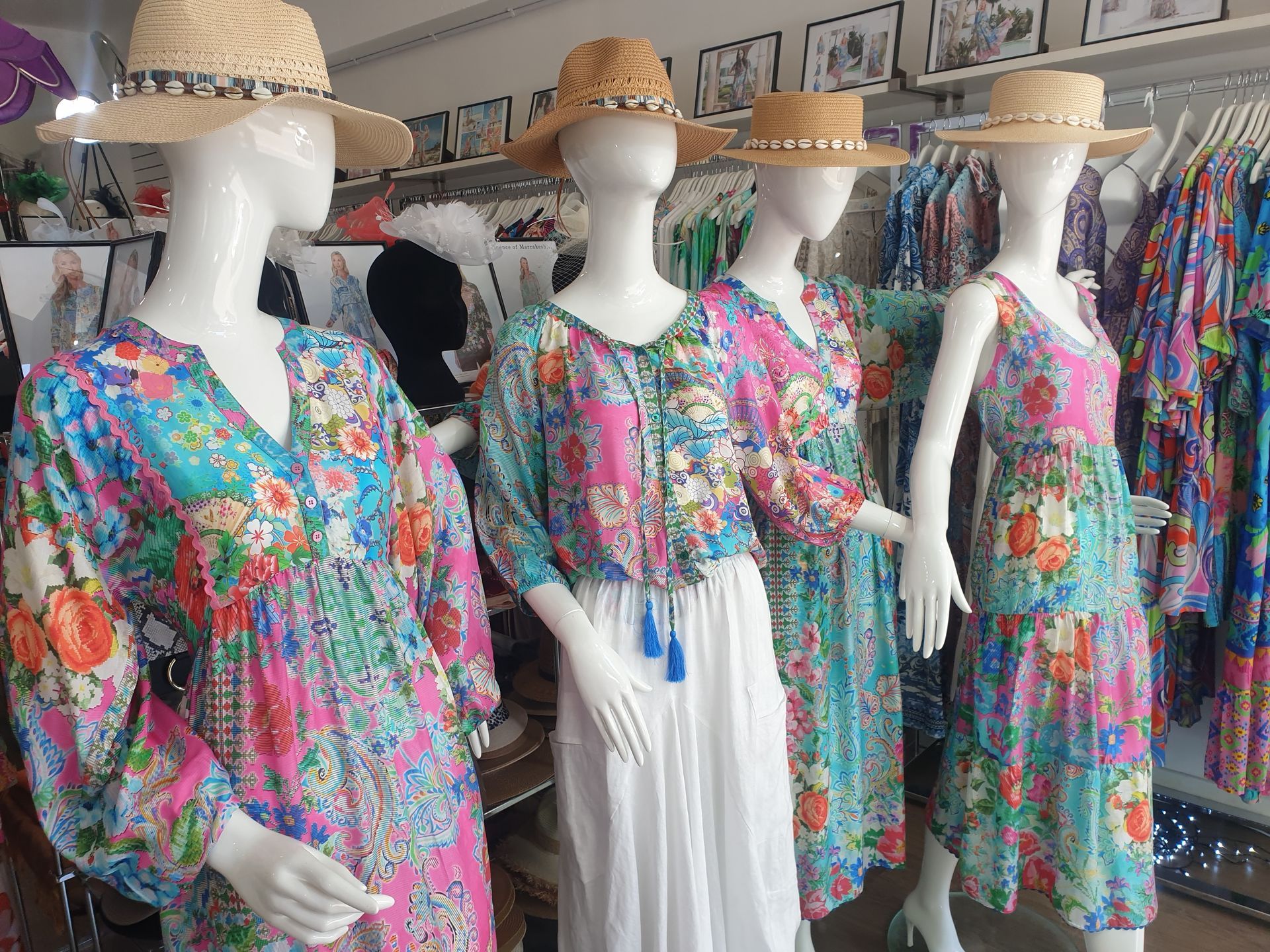 Four mannequins in a clothing store wearing floral dresses and straw hats; clothes racks in the background.