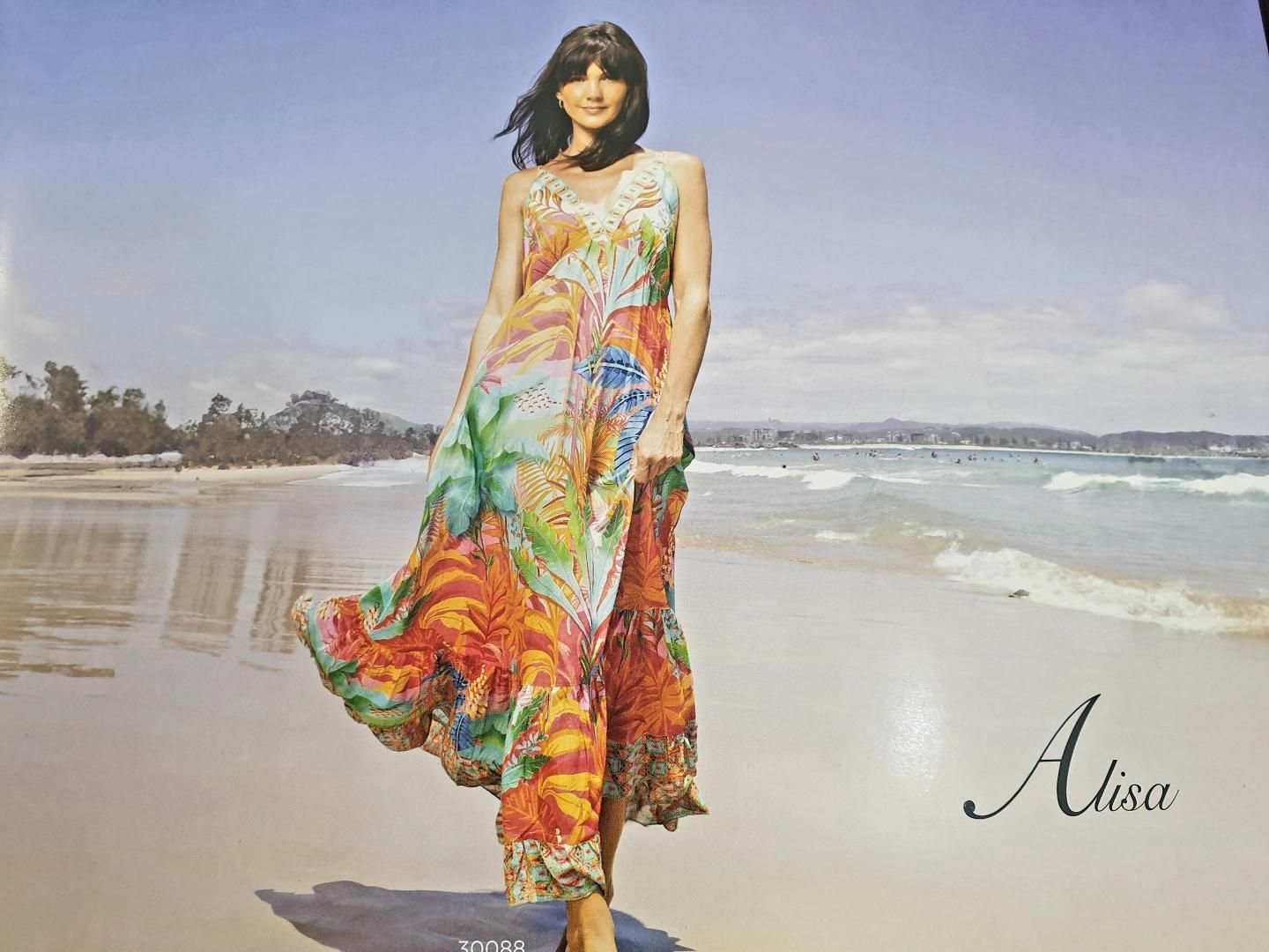 Woman in colorful dress on a beach, smiling. Sunny day, waves in background.