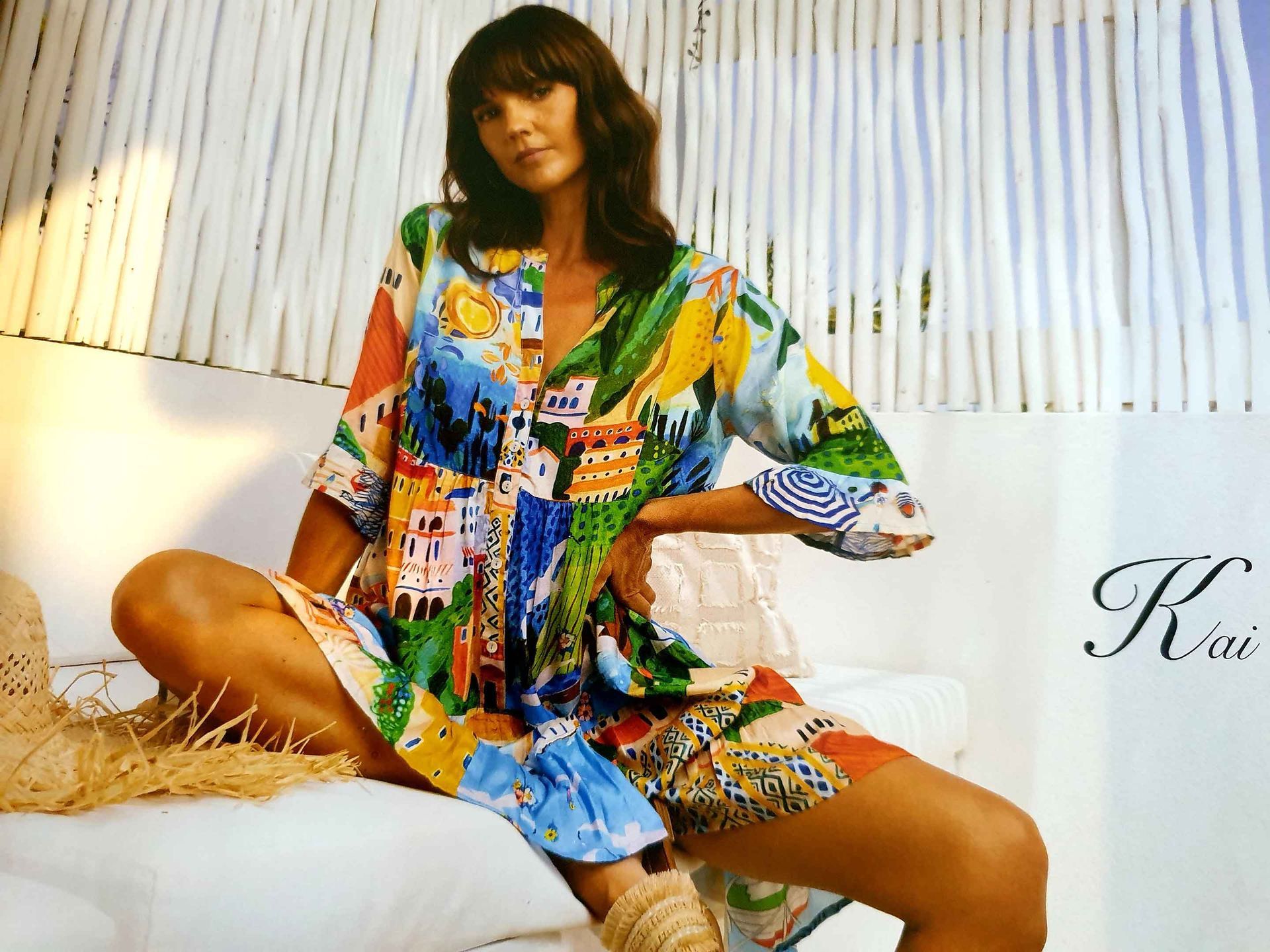 Woman in colorful print dress sits, hand on hip, on white surface; neutral setting.