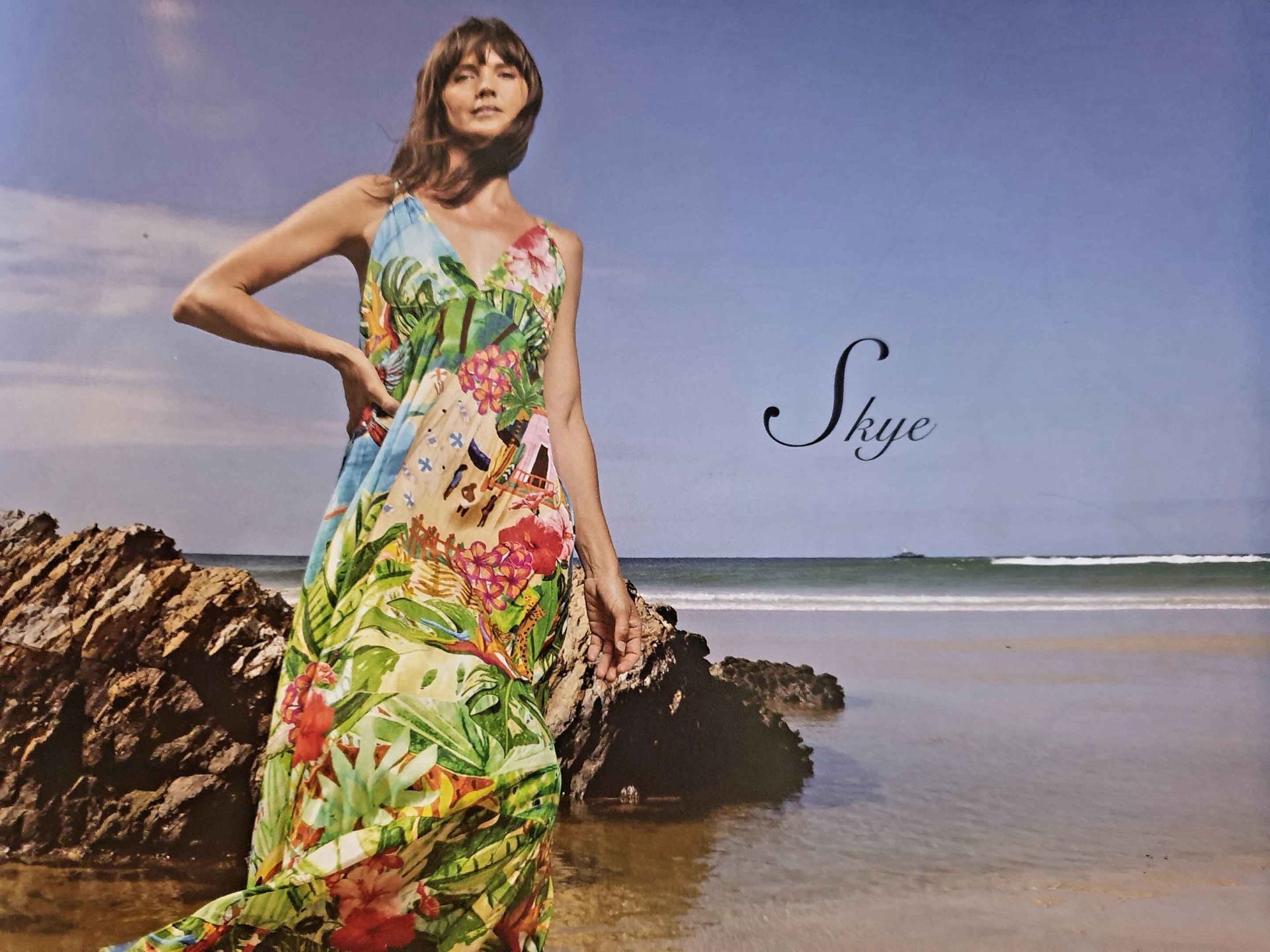 Woman in floral dress on a beach, hand on hip, looking towards the viewer. Sky and ocean in background.