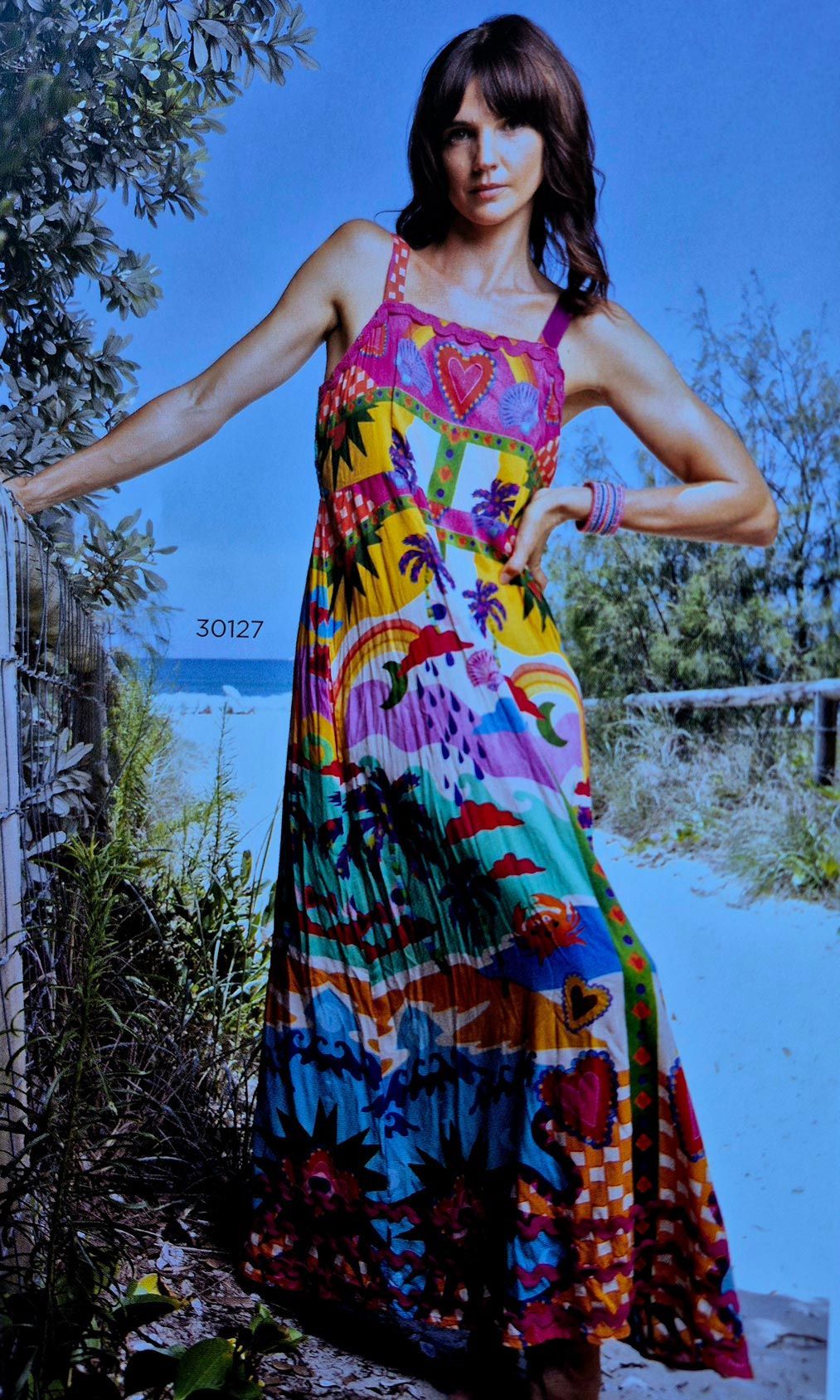 Woman in a colorful floral maxi dress poses outdoors, leaning on a gate near a beach.