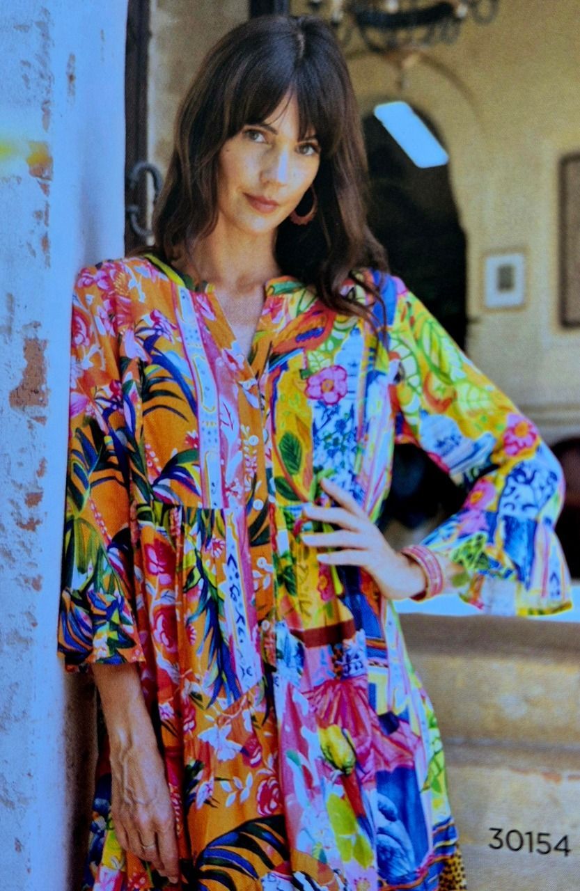 Woman in colorful floral dress, leaning against a wall, hand on hip, looking at the camera.