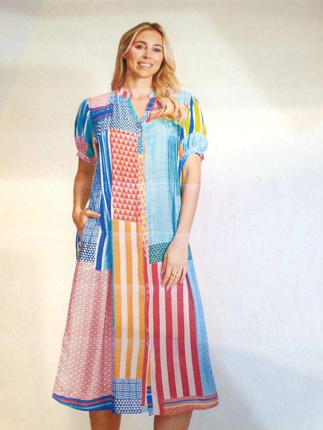 Woman in colorful patchwork dress smiles, standing indoors with hands in pockets.