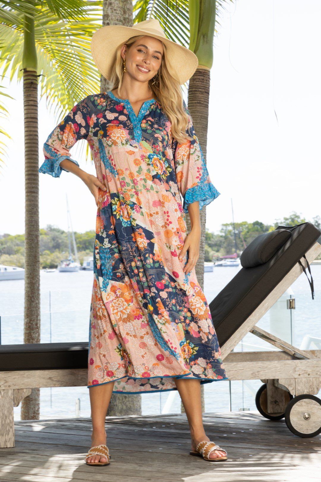 Woman in floral dress and straw hat smiles, standing near waterfront, wearing sandals.