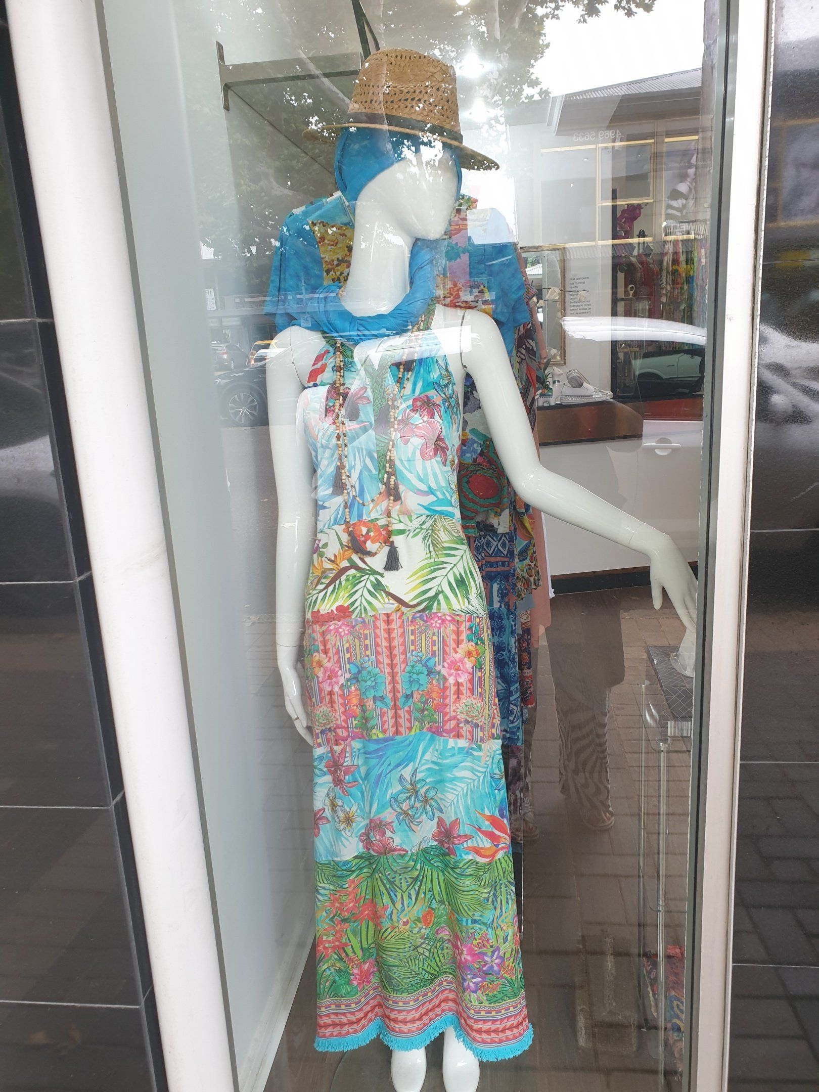 Mannequin in a colorful floral maxi dress in a store window, wearing a blue scarf and hat.