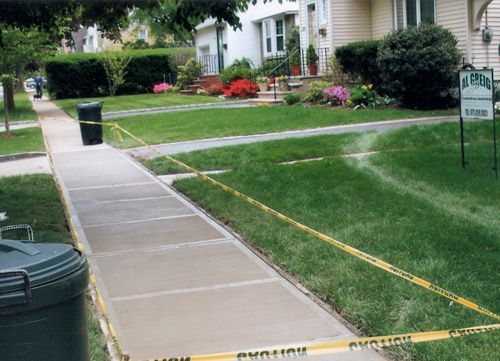 Newly poured concrete sidewalk blocked by caution tape on a residential street. Green grass and houses in the background.