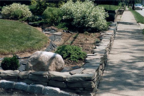 Stone retaining wall bordering a sidewalk and garden with greenery and a large rock.
