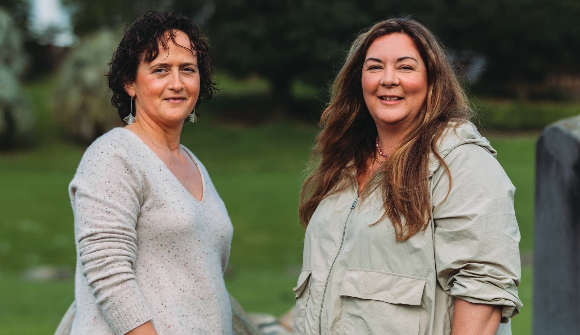 Two women standing outdoors, smiling at the camera in a green park setting.