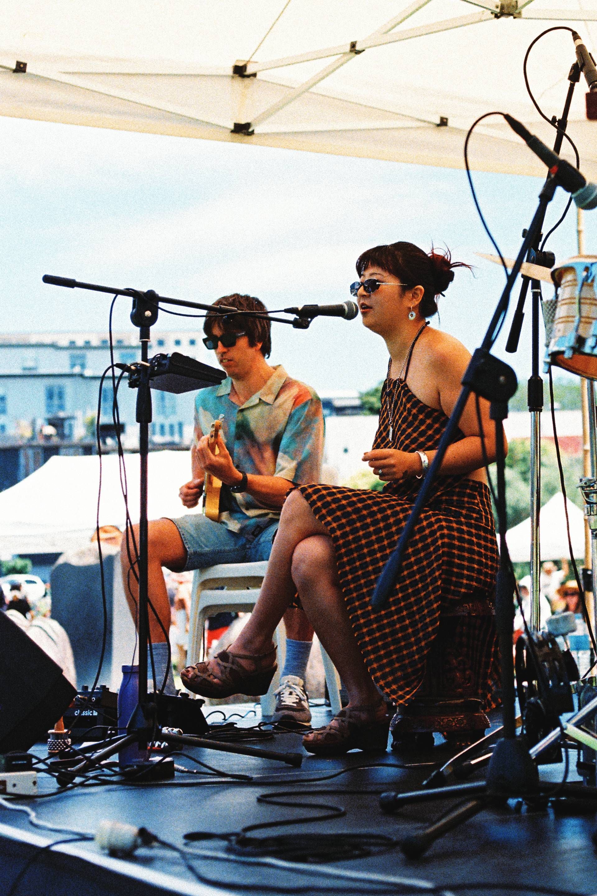 Two musicians performing under a white canopy, seated with microphones and acoustic guitars.