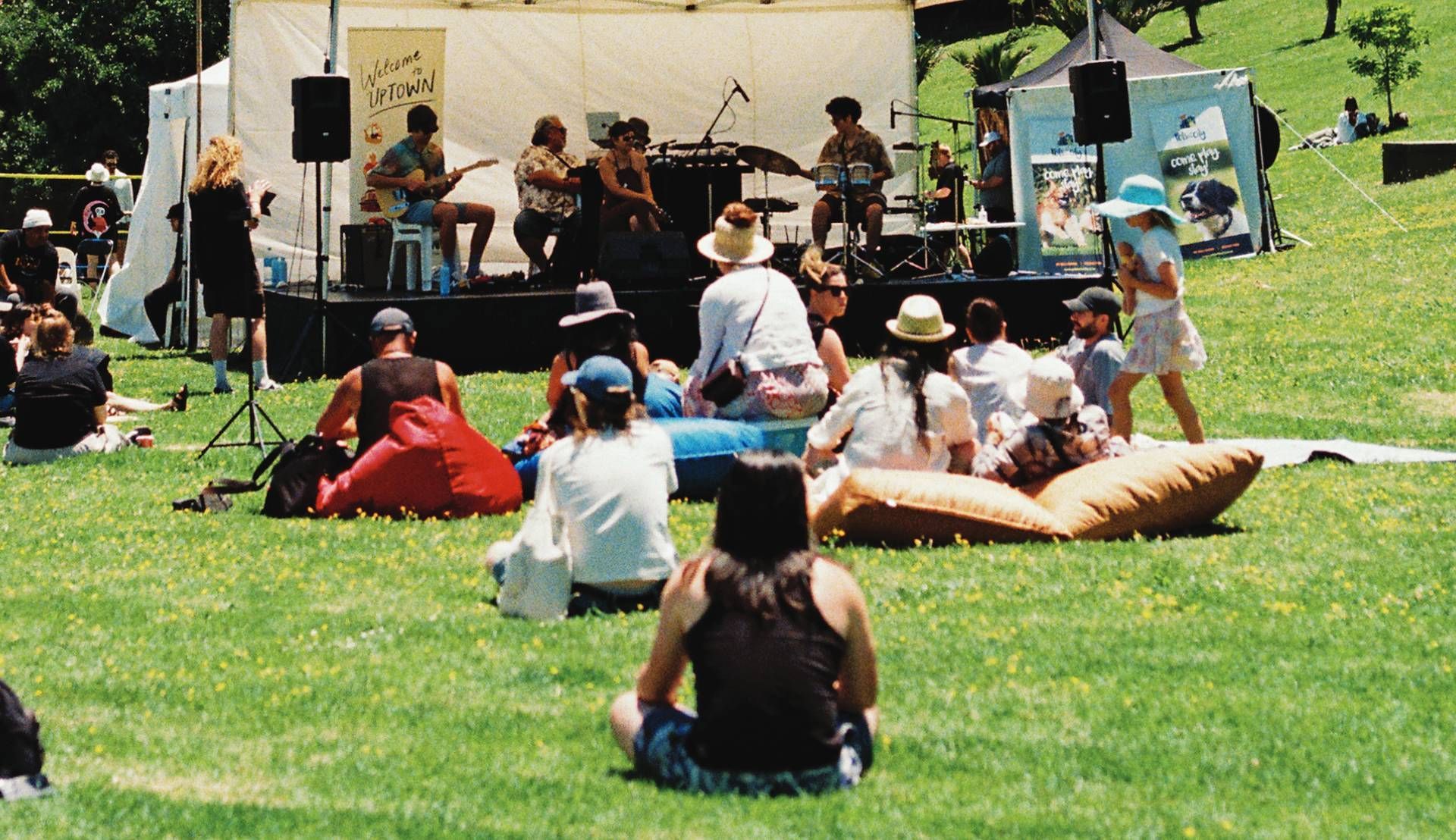 People relaxing on a sunny grassy lawn near a white event tent with a small stage and crowd.