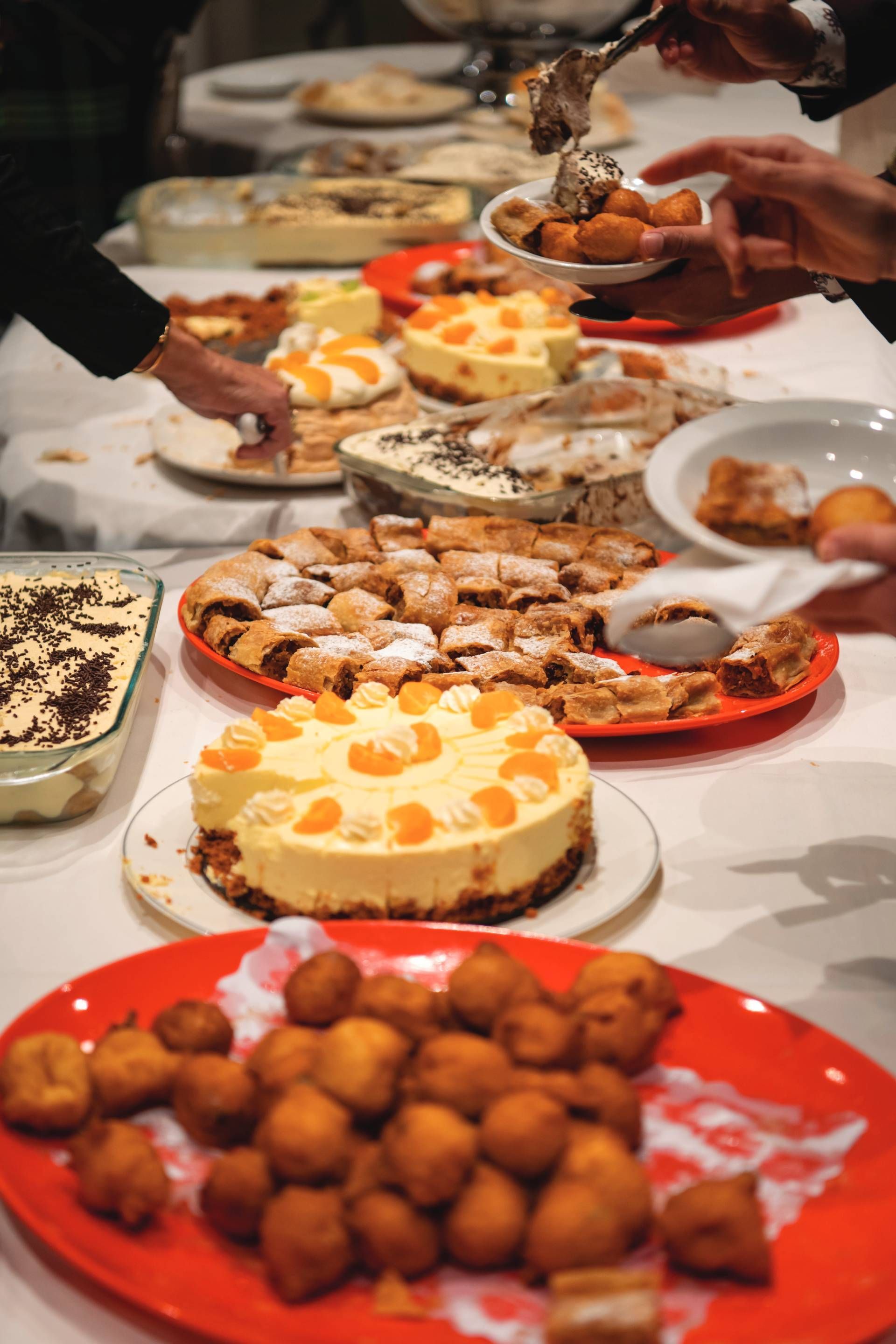 Dessert buffet with cheesecake, pastries, and fruit on a table as hands serve food.