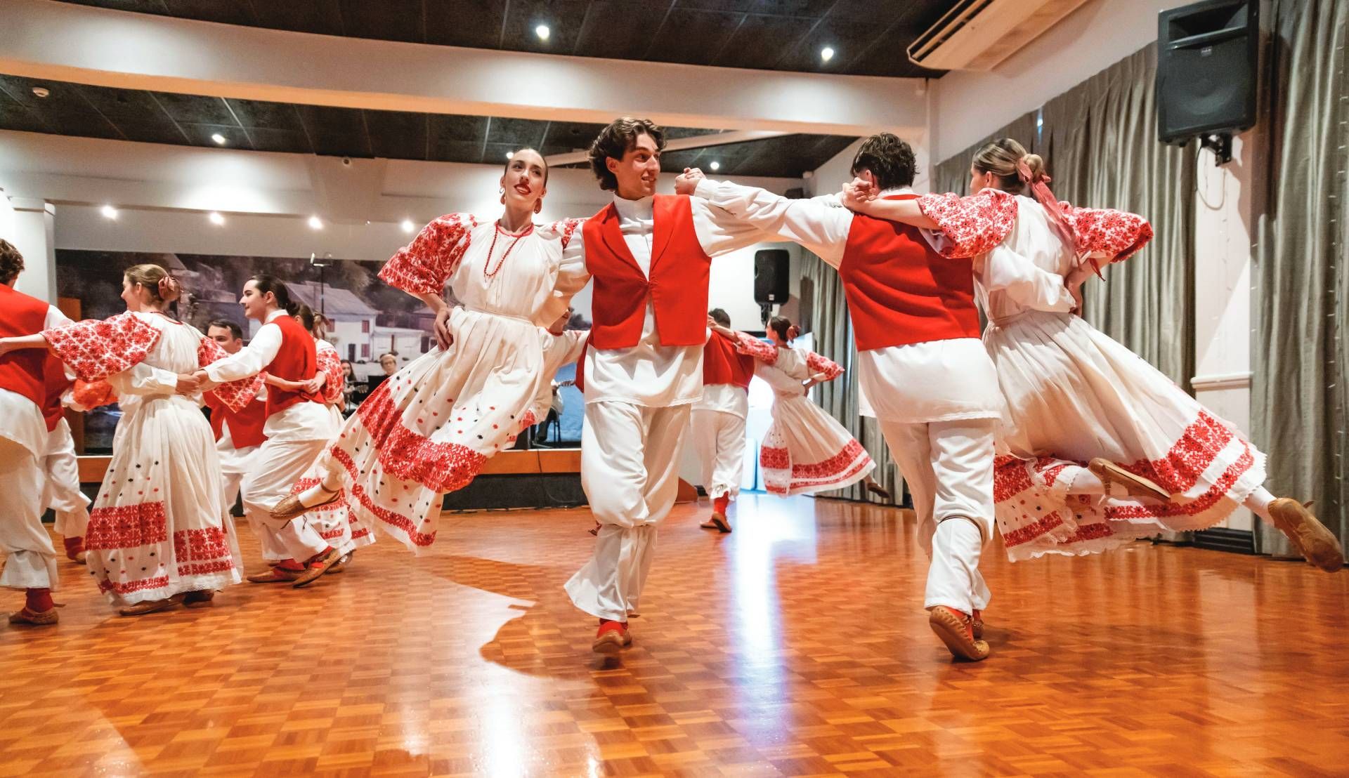 Dancers in red and white costumes perform a traditional folk dance in a wooden hall.
