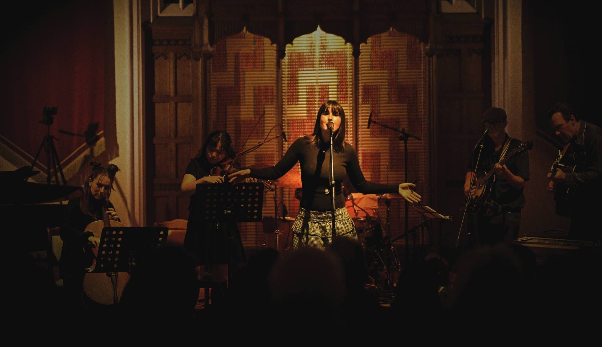 Band performing on stage under warm orange lighting, with a singer centered before a glowing backdrop.