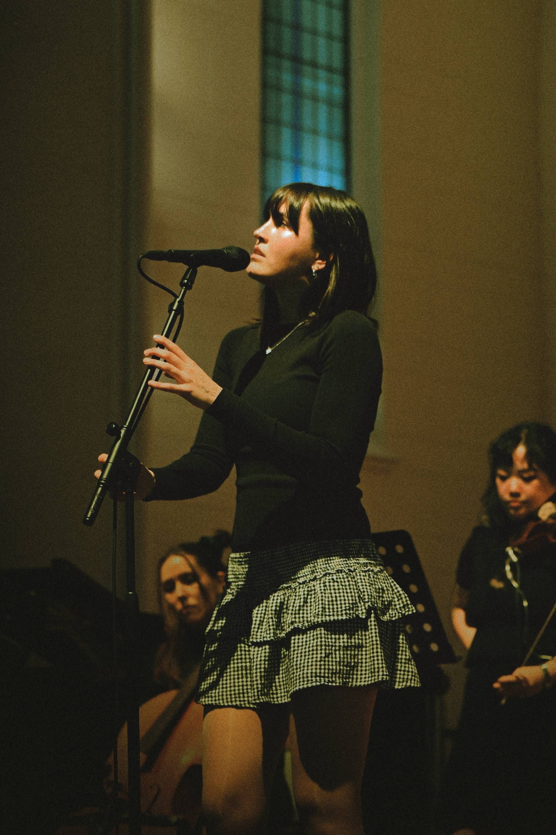 Singer at a microphone onstage, wearing a black top and plaid skirt, with band members behind her.