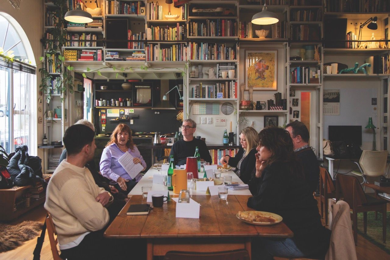 People seated around a wooden table in a room filled with bookshelves, bottles, and food.