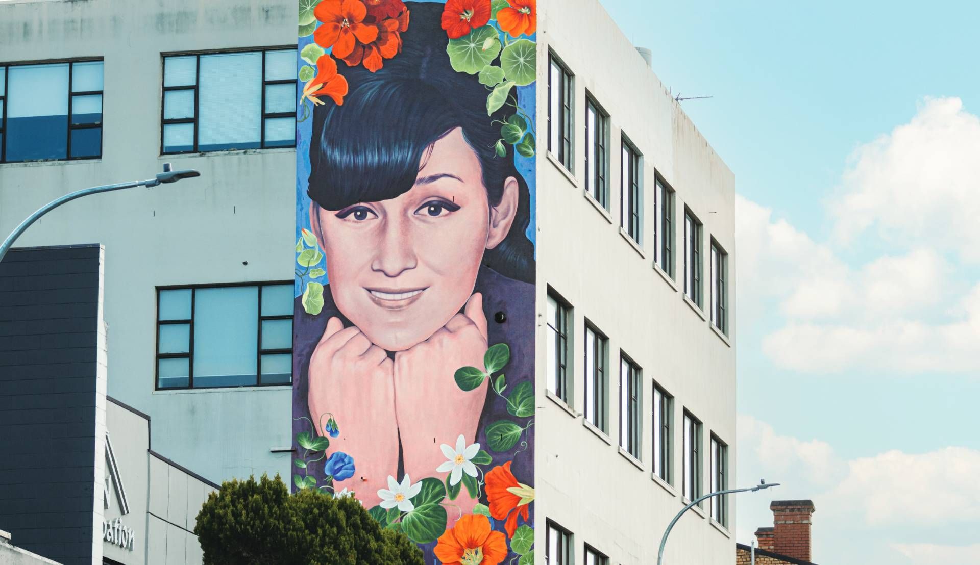 Colorful mural of a person’s face on a white building beside windows, with flowers and blue sky.
