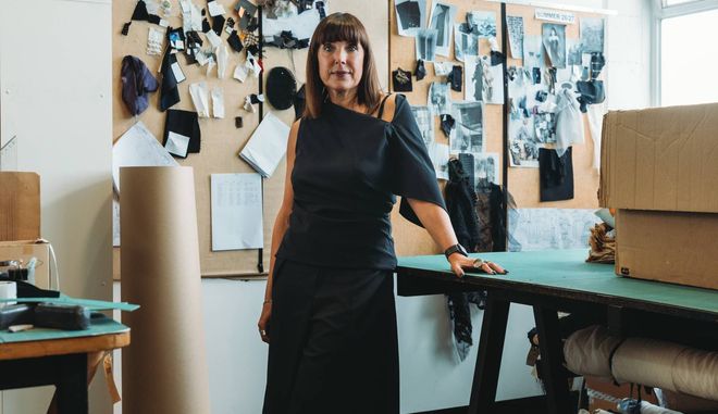 Woman in black dress standing in a studio, leaning on a table beside pinned photos and stacked boxes.