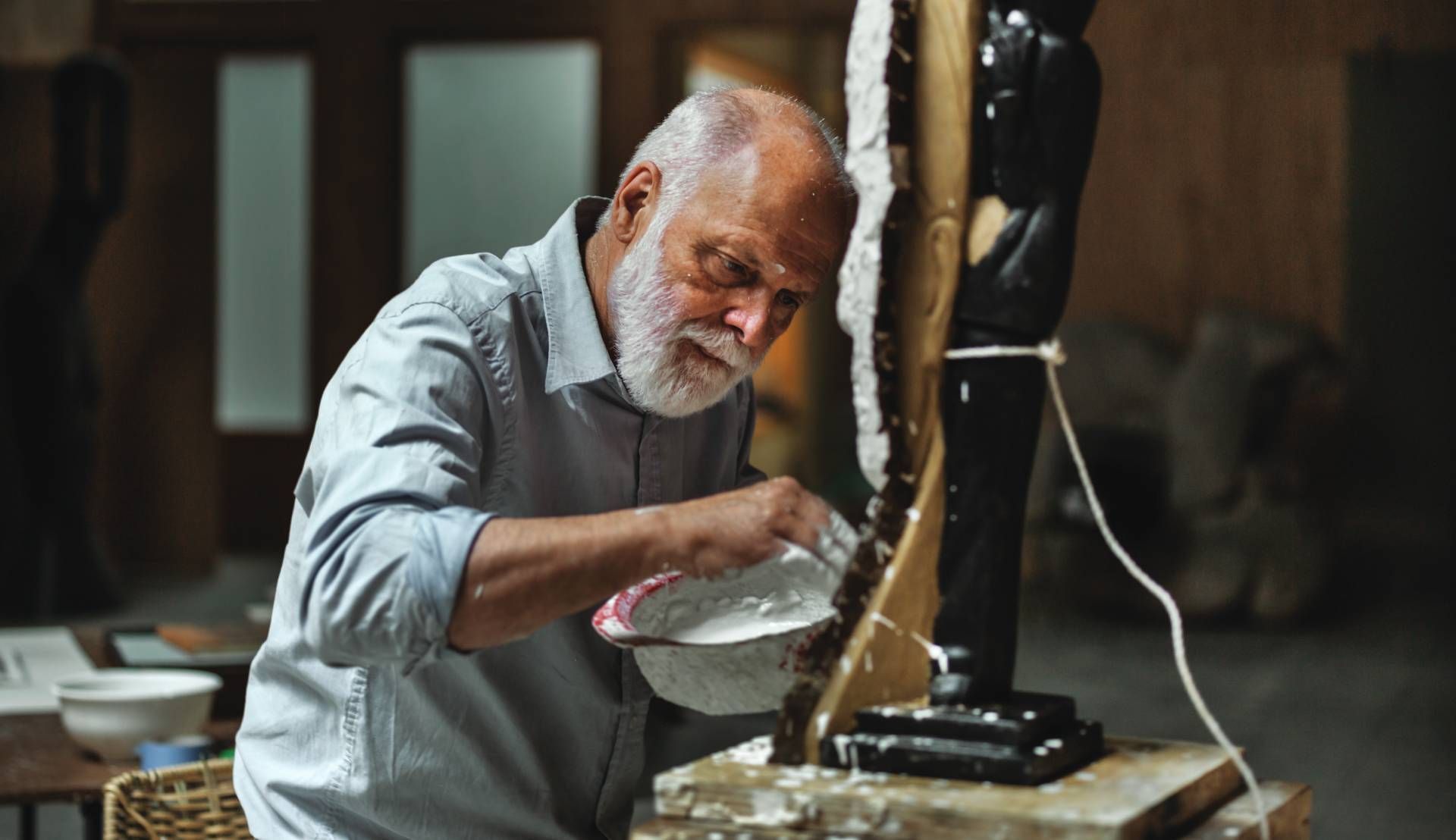 Elderly man in a workshop, pouring liquid into a mold beside a vertical machine.