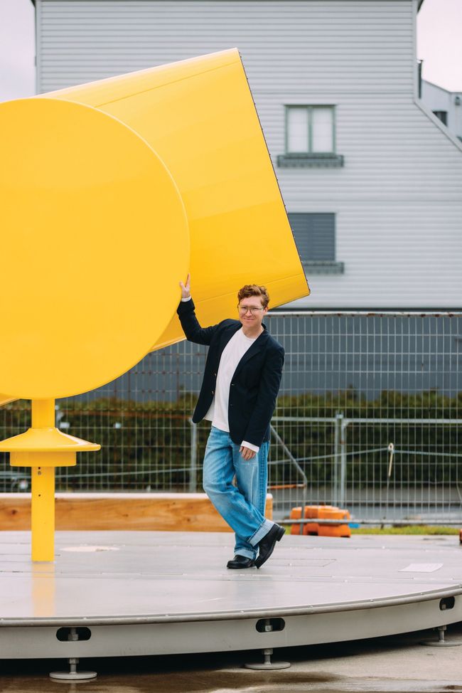 Man standing beside a large yellow sculpture on a circular platform outdoors