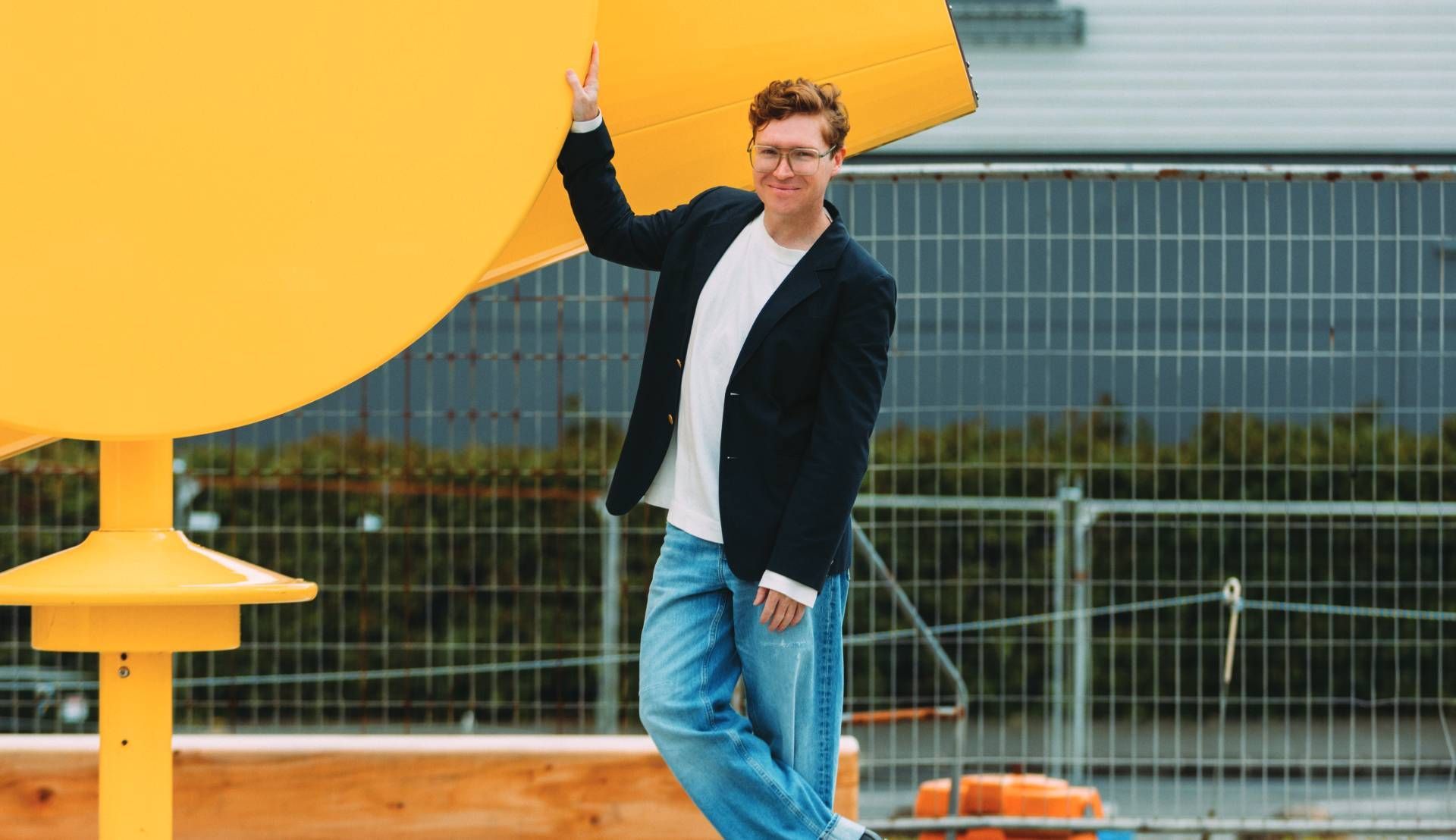 Person standing outdoors beside a large yellow structure, waving in front of a fenced building.