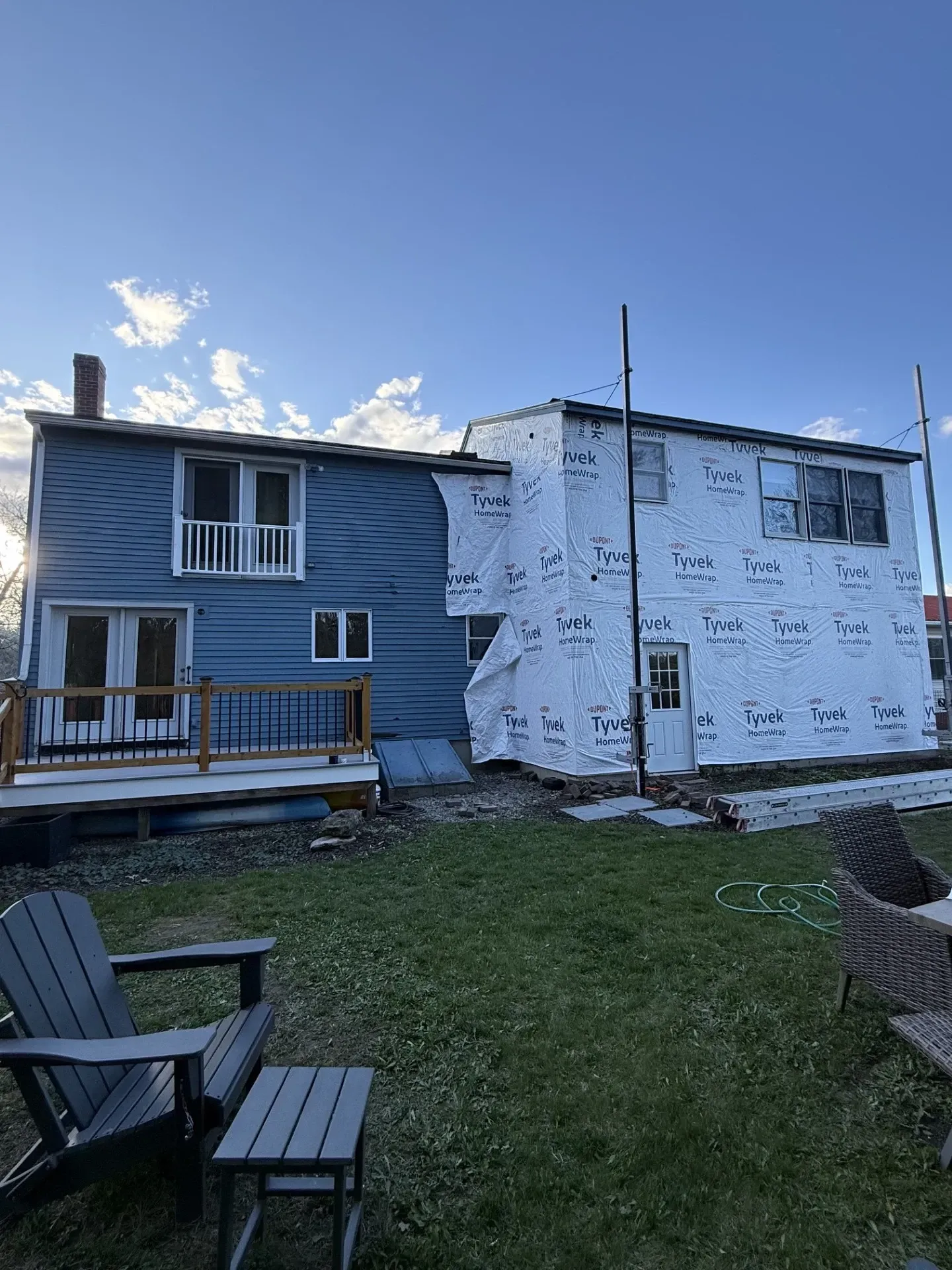 Two-story house under renovation with white wrap on one side and a small wooden deck in a grassy yard.
