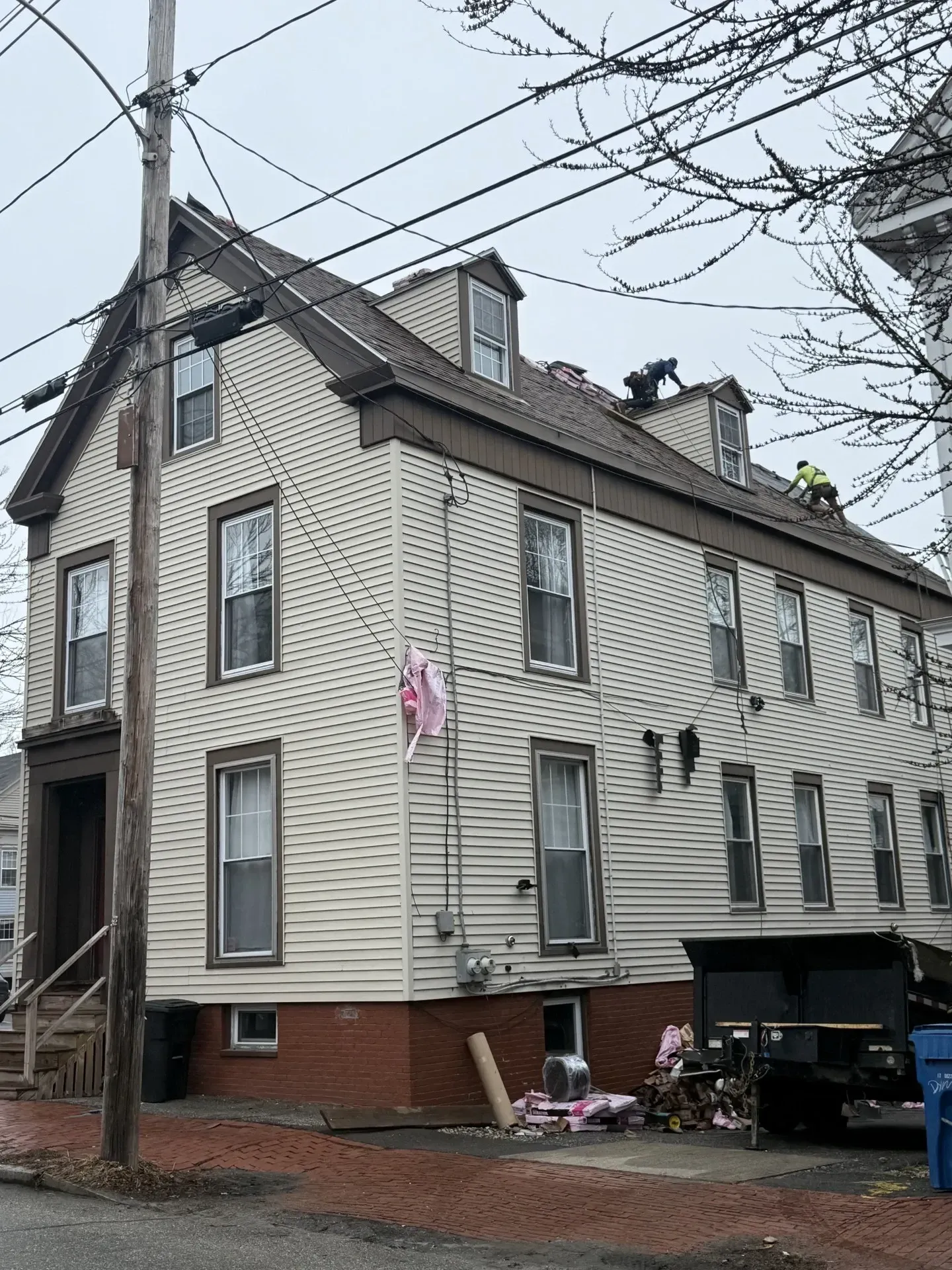 Three-story gray house with brick base, dormer windows, and a driveway under overcast sky.