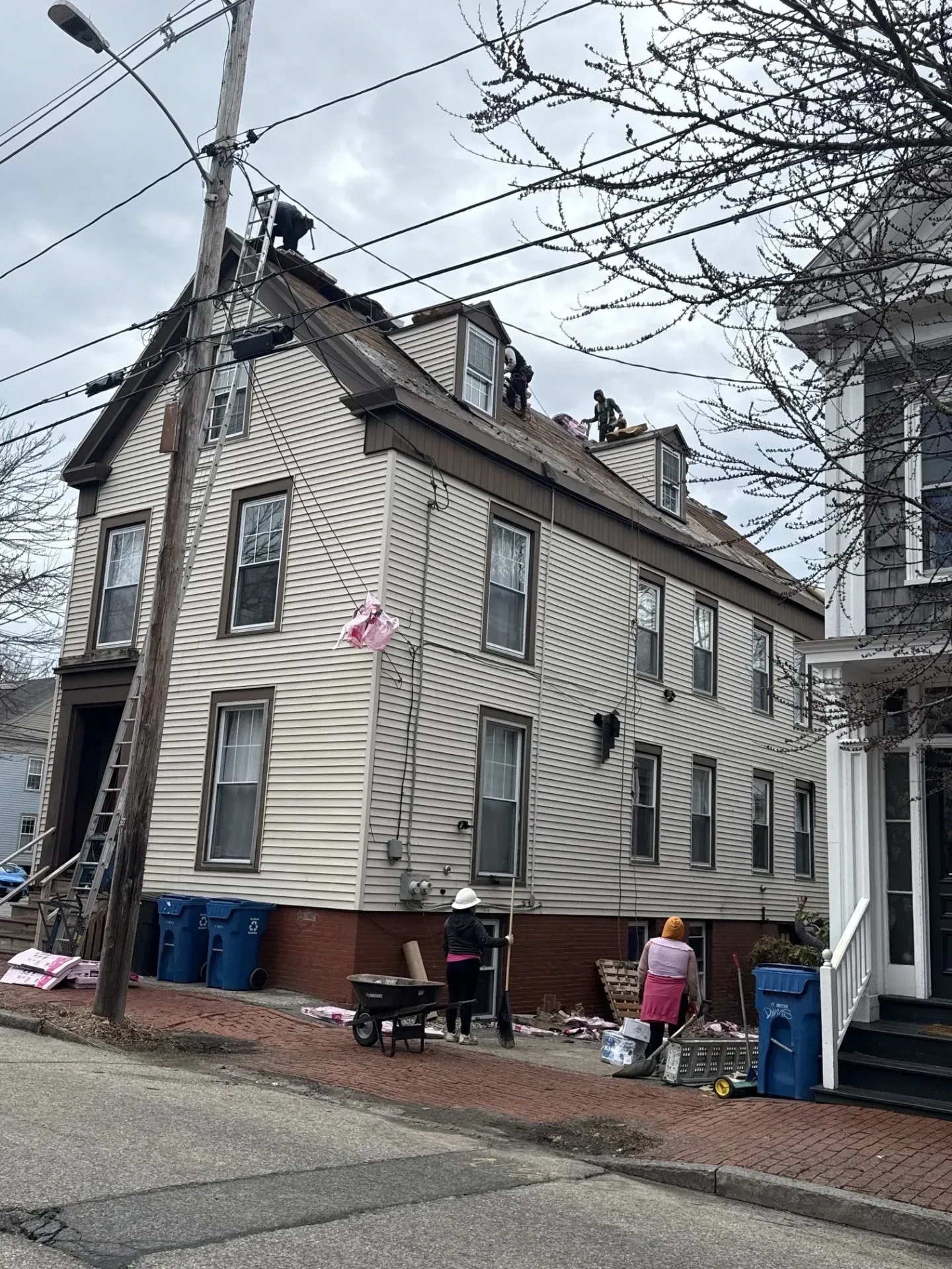 Three-story gray house with boarded windows and people by trash bins on a residential street