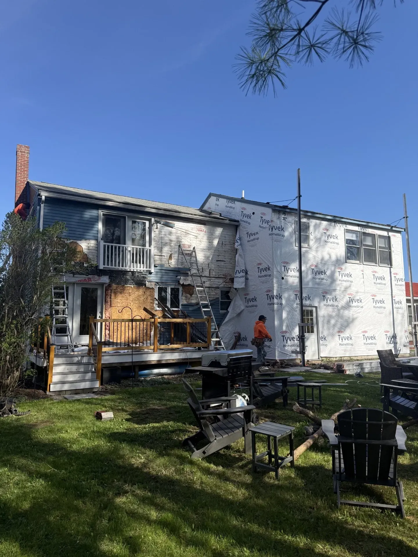 House exterior under renovation with workers, ladders, and tools in a grassy yard under a clear blue sky